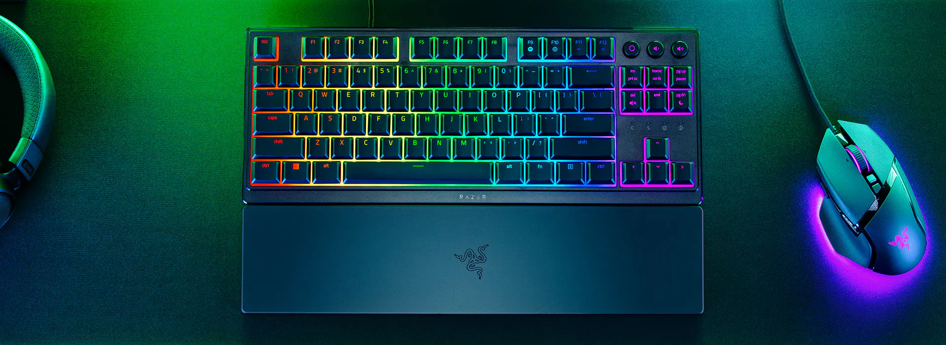 Fekete Razer billentyűzet szivárványos háttérvilágítású billentyűkkel, hozzáillő csuklótámasszal, egérrel és fejhallgatóval.