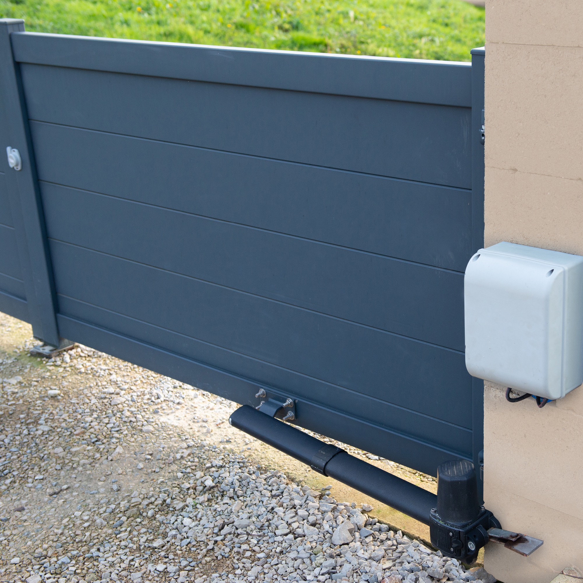Ein graues Schiebetor ist teilweise geöffnet. Eine weiße Box und ein schwarzer Motor sind an einer beigen Wand befestigt. Kiesboden.
