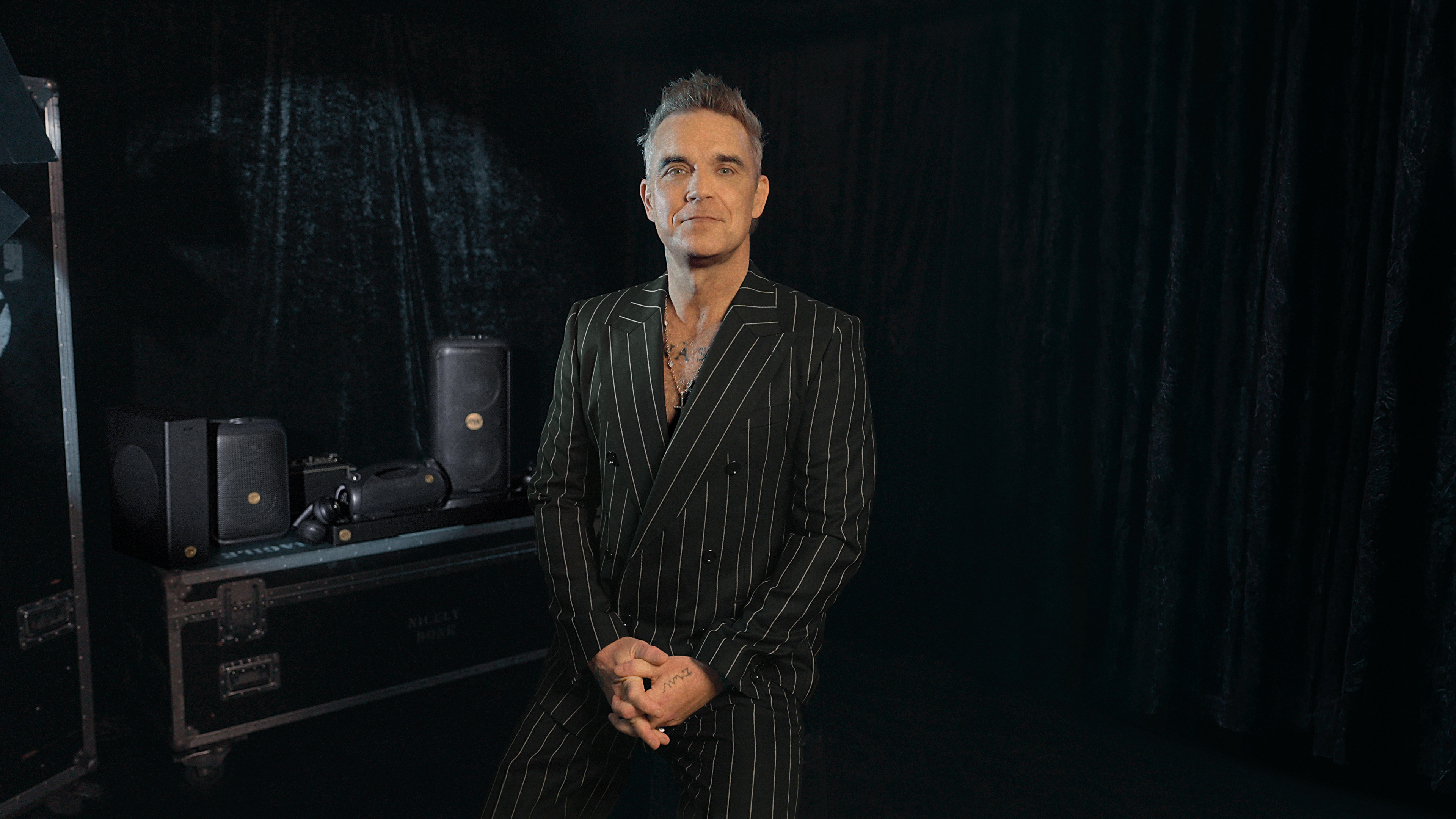 Mężczyzna w pasiastym garniturze stoi przed ciemną zasłoną. Sprzęt sceniczny po lewej. Ma tatuaże i naszyjnik.