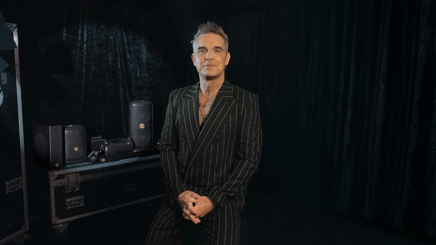 Mężczyzna w czarnym garniturze w paski, splecione dłonie. Głośniki i sprzęt po lewej, ciemne tło.