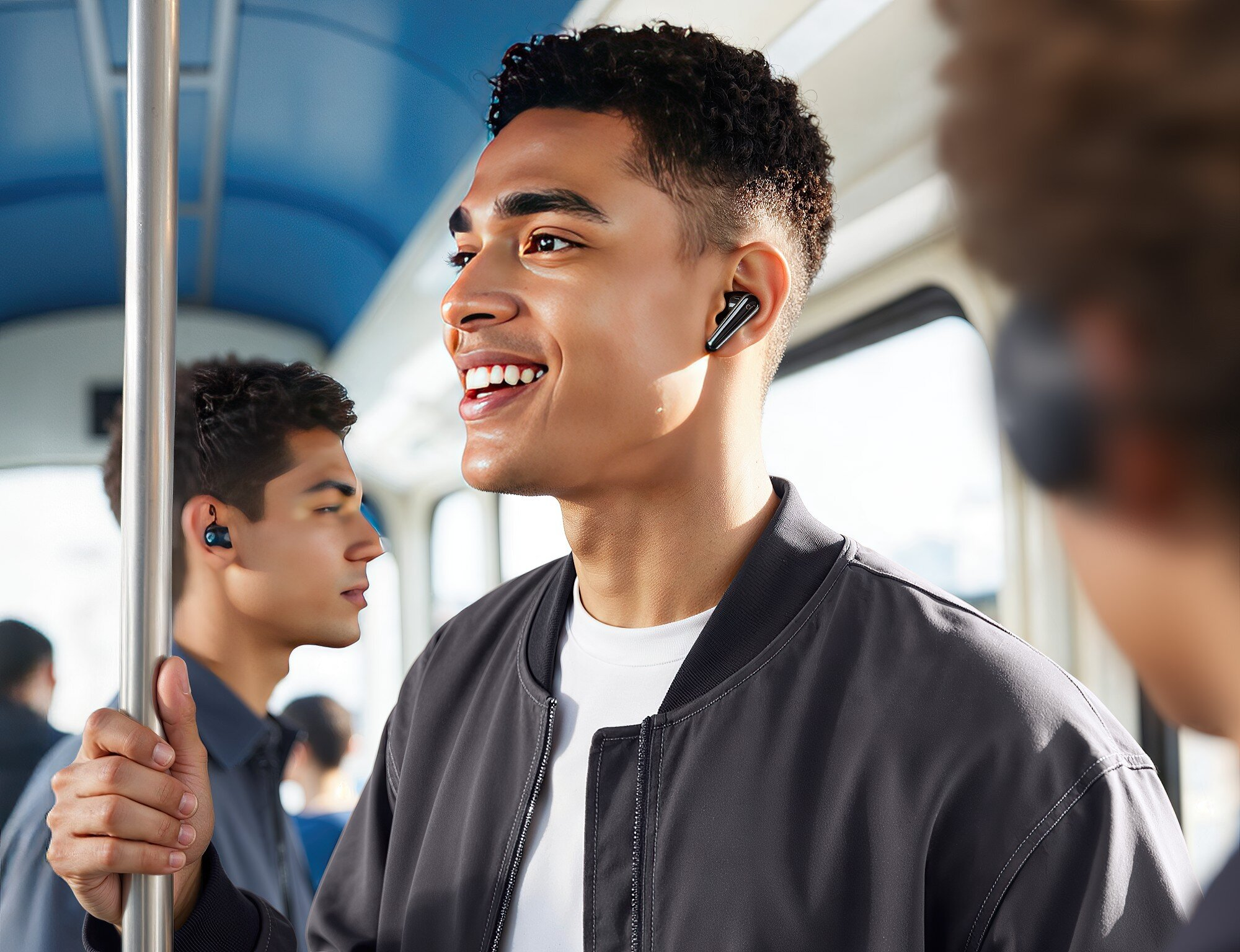 Uśmiechnięty mężczyzna w słuchawkach w pociągu. Dwóch innych mężczyzn w tle. Mężczyzna ma na sobie szarą kurtkę i białą koszulę.