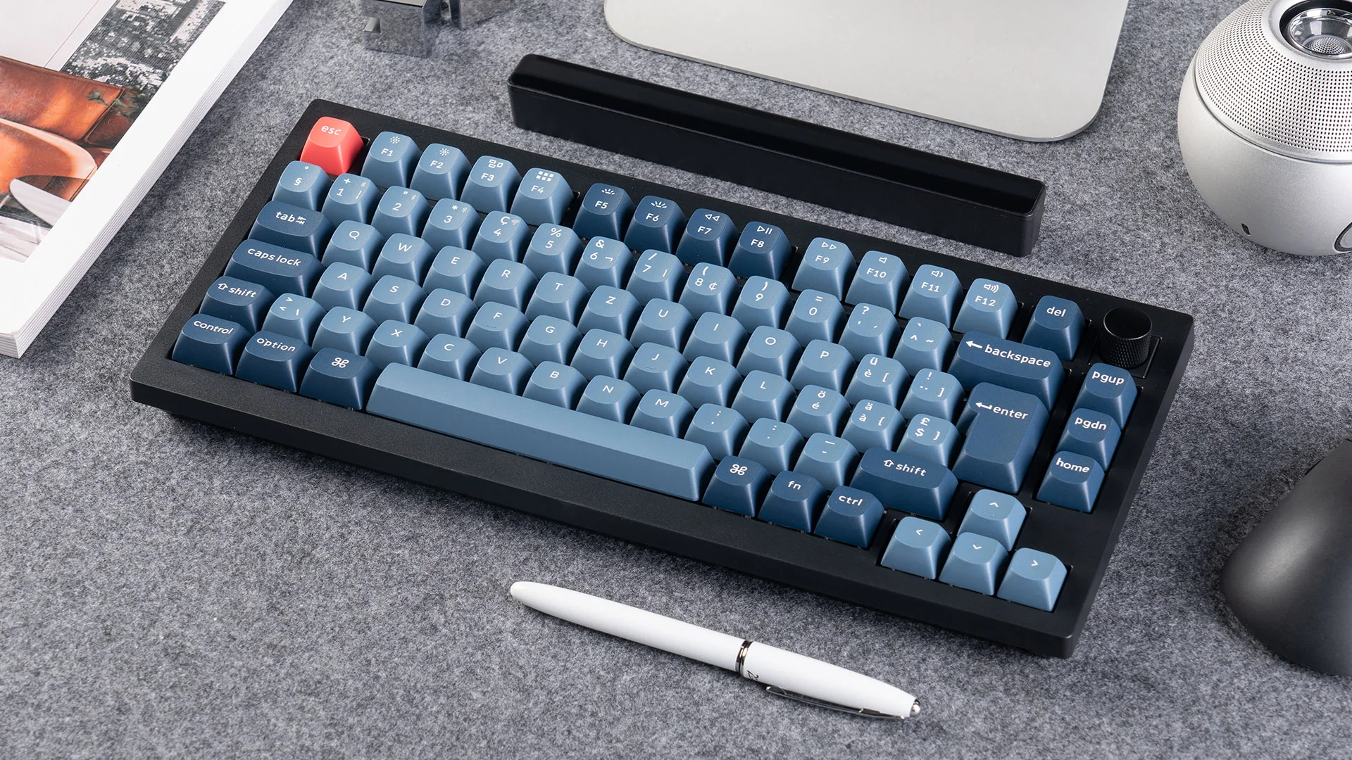 Un clavier noir avec des touches bleues et une touche Échap rouge est sur une surface grise, à côté d'un stylo, d'une souris et d'un haut-parleur.