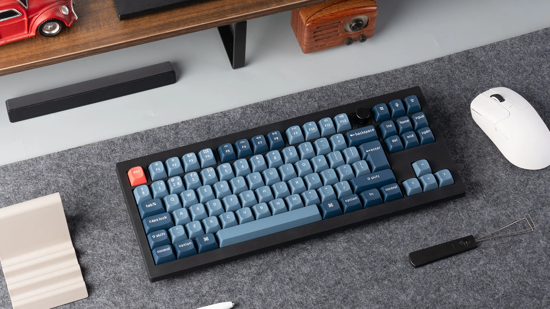 Eine schwarze mechanische Tastatur mit blauen Tasten und einer roten Escape-Taste liegt auf einer grauen Oberfläche. Rechts befindet sich eine weiße Maus.