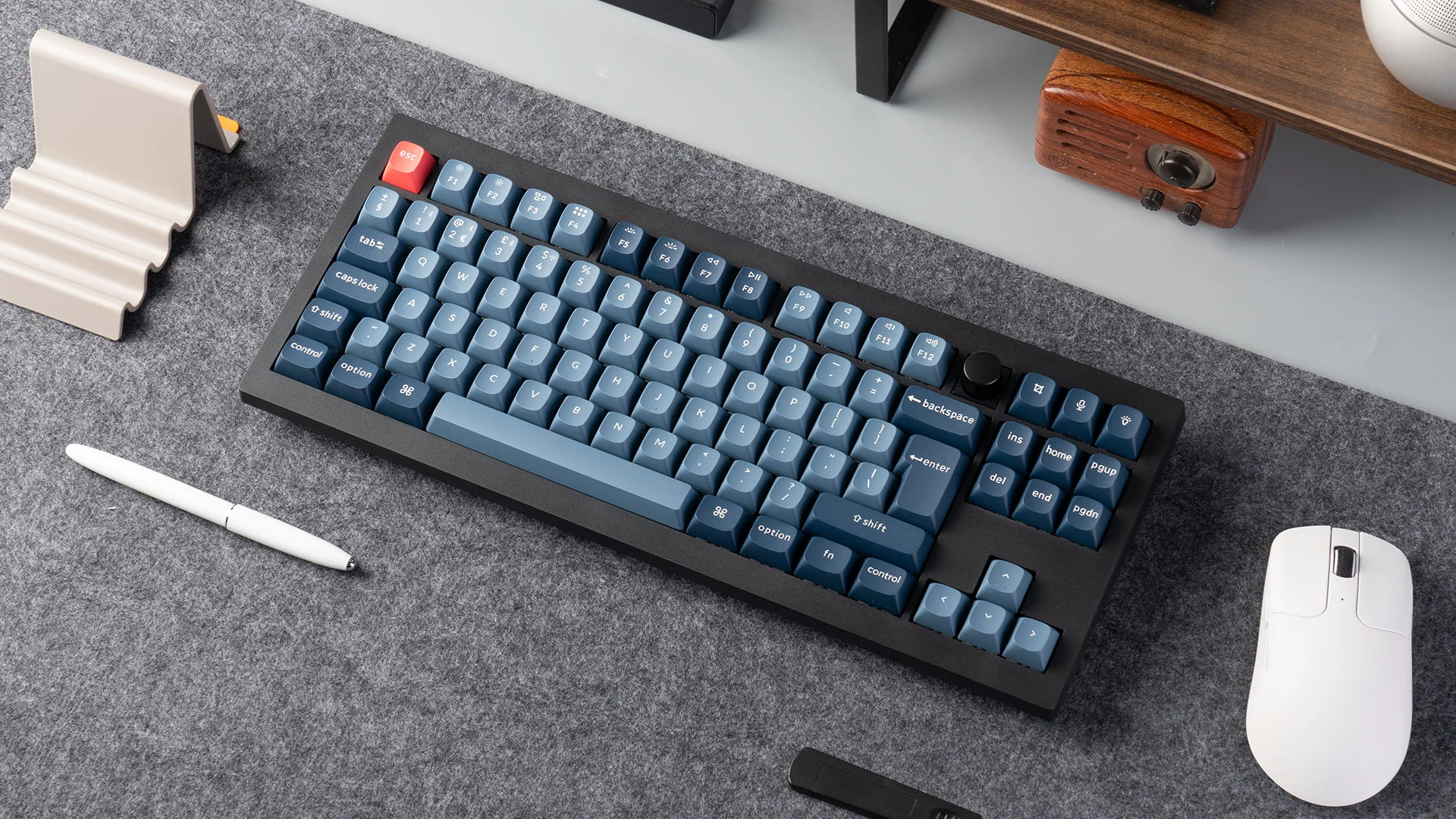 Eine Tastatur mit blauen Tasten, eine rote Esc-Taste. Eine weiße Maus und ein Stift liegen auf einer grauen Oberfläche. Ein Holzradio ist im Hintergrund.