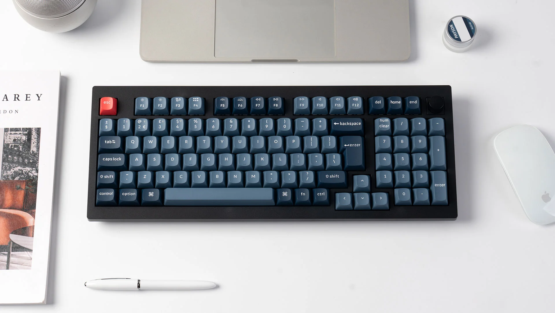 Draufsicht auf eine Tastatur mit blauen Tasten und einer roten Escape-Taste, mit Laptop, Maus und Stift auf weißer Oberfläche.