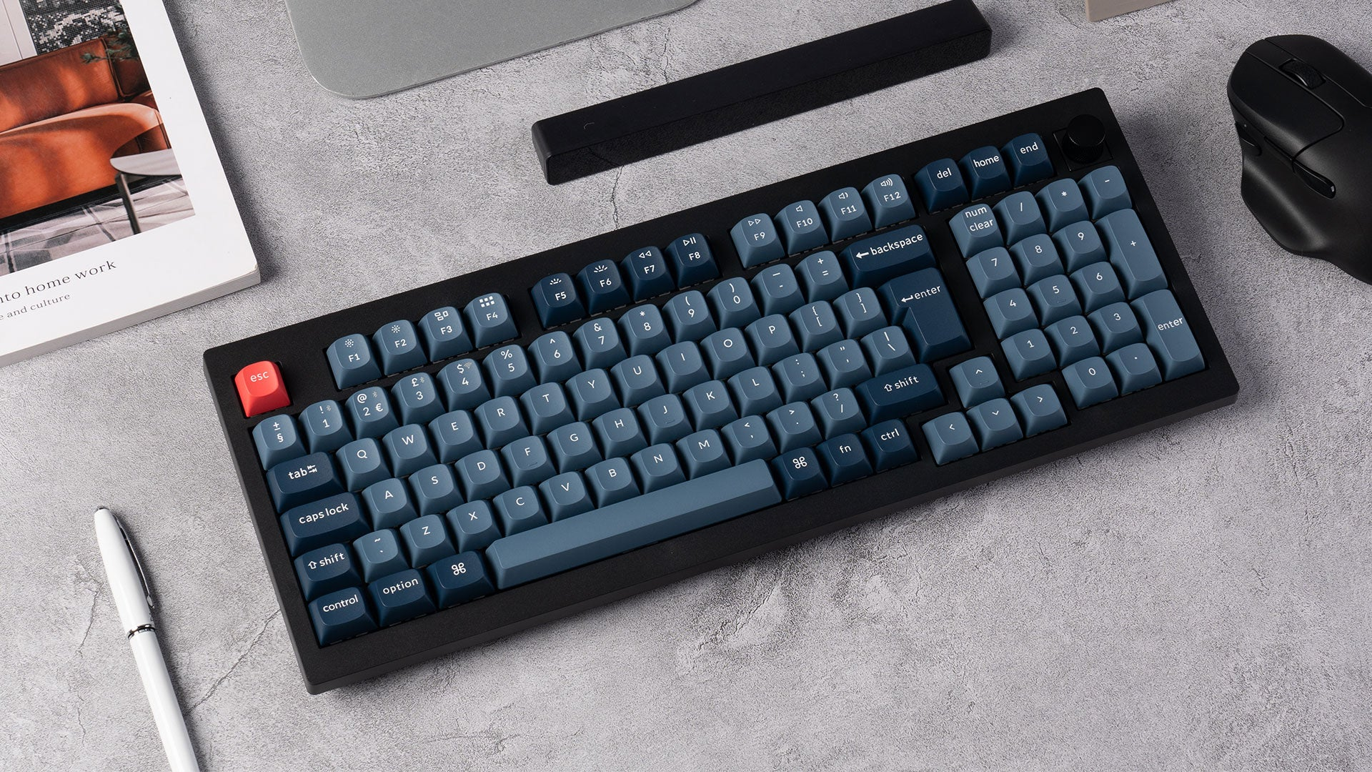 Eine schwarze Tastatur mit blauen Tasten, eine rote Escape-Taste, eine Maus und ein Buch liegen auf grauem Untergrund.
