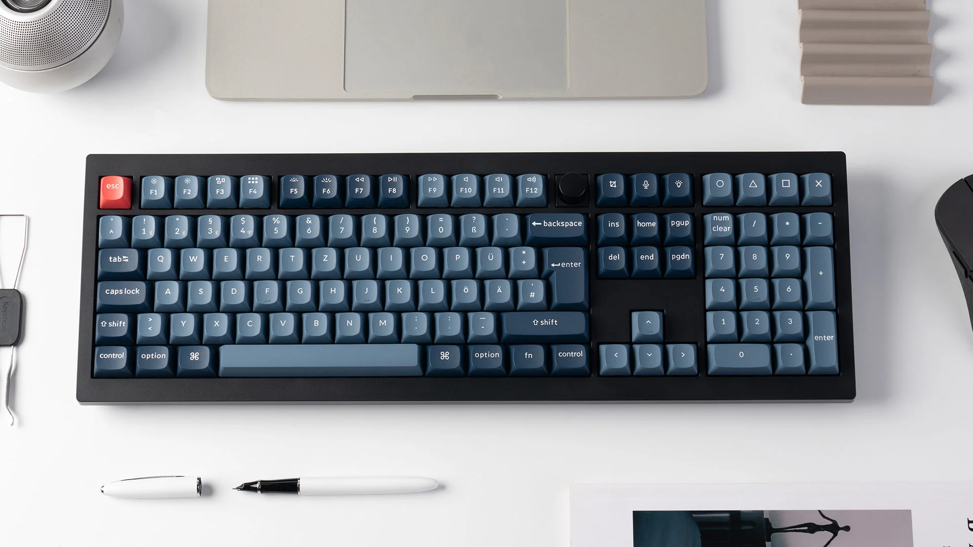 Eine schwarze Keychron-Tastatur mit blauen Tasten, roter Escape-Taste, weißem Stift und Laptop auf weißer Oberfläche.