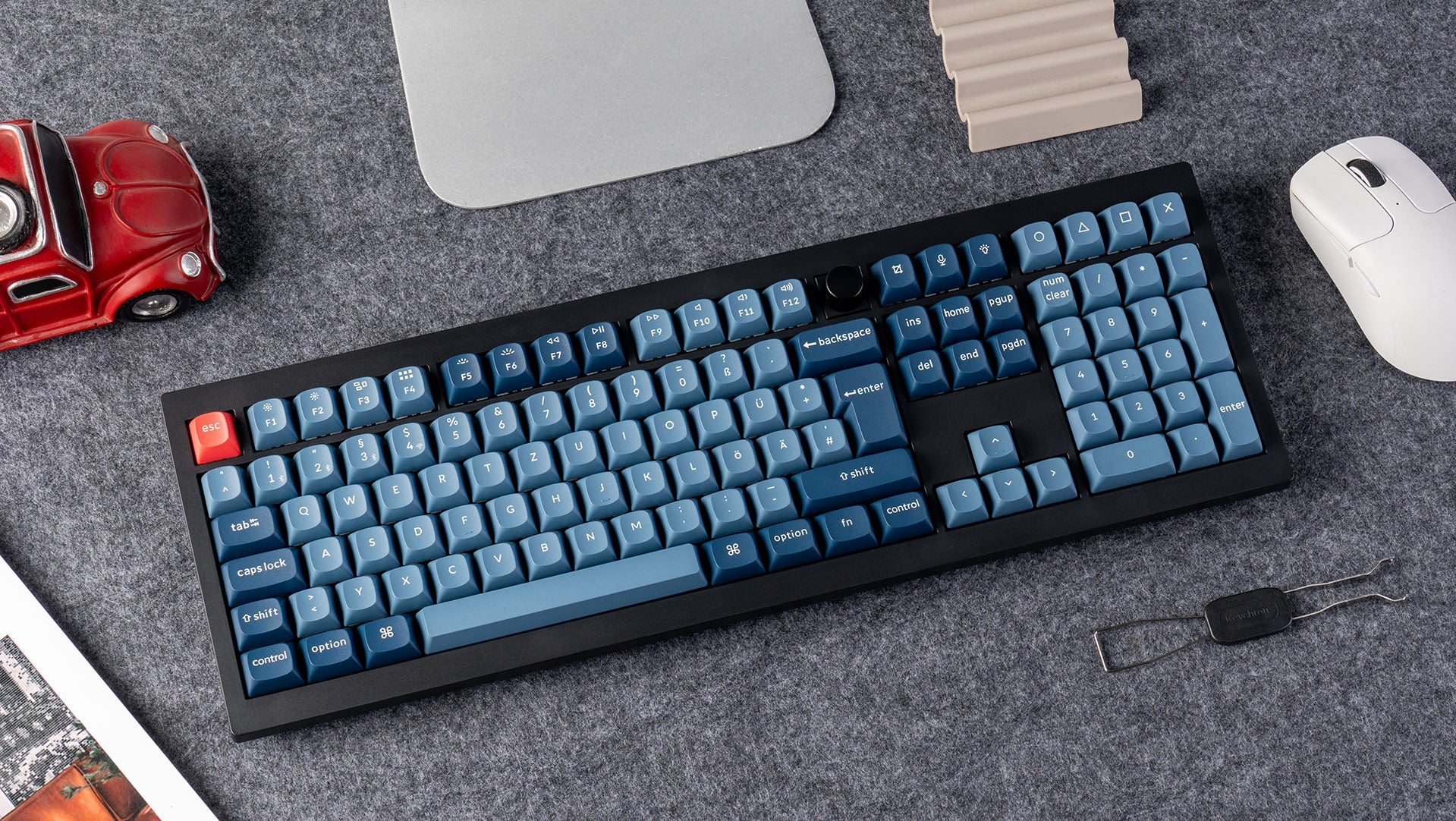Mechanische Tastatur mit blauen Tasten, rotes Spielzeugauto, weiße Maus und grauer Hintergrund.