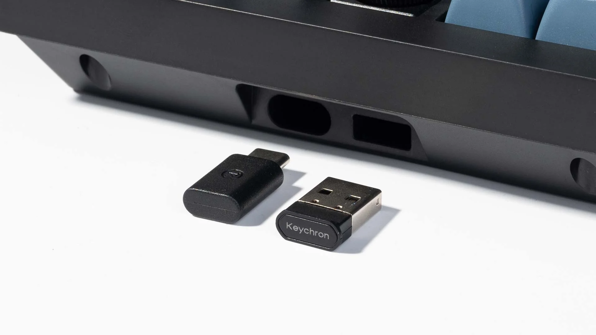 Ein schwarzer Tastatursockel mit USB-C-Adapter und USB-Empfänger. Der Empfänger hat das Keychron-Logo.
