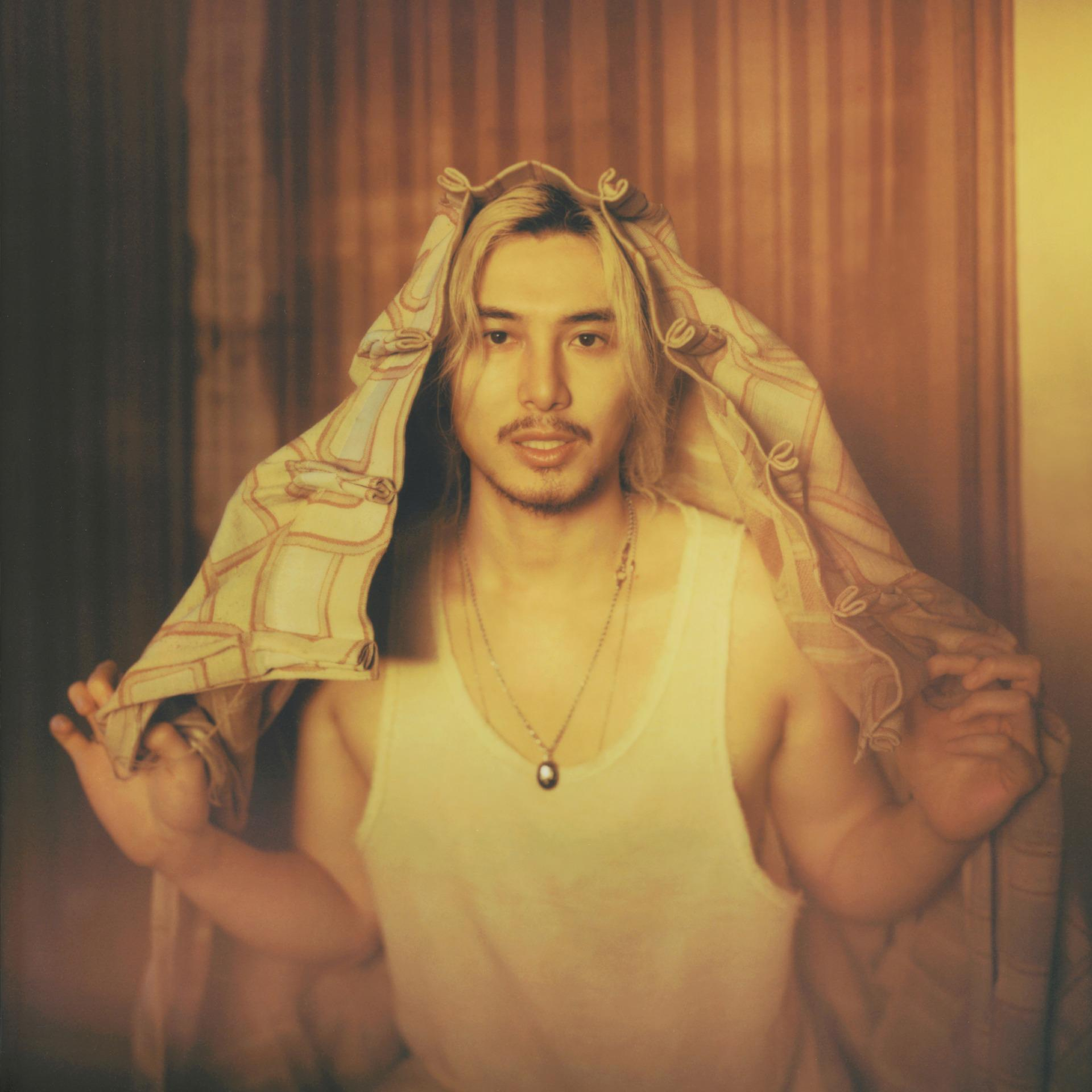 Ein Mann mit blonden Haaren, der ein weißes Tanktop trägt und ein gemustertes Tuch und eine Halskette hält. Brauner Hintergrund.