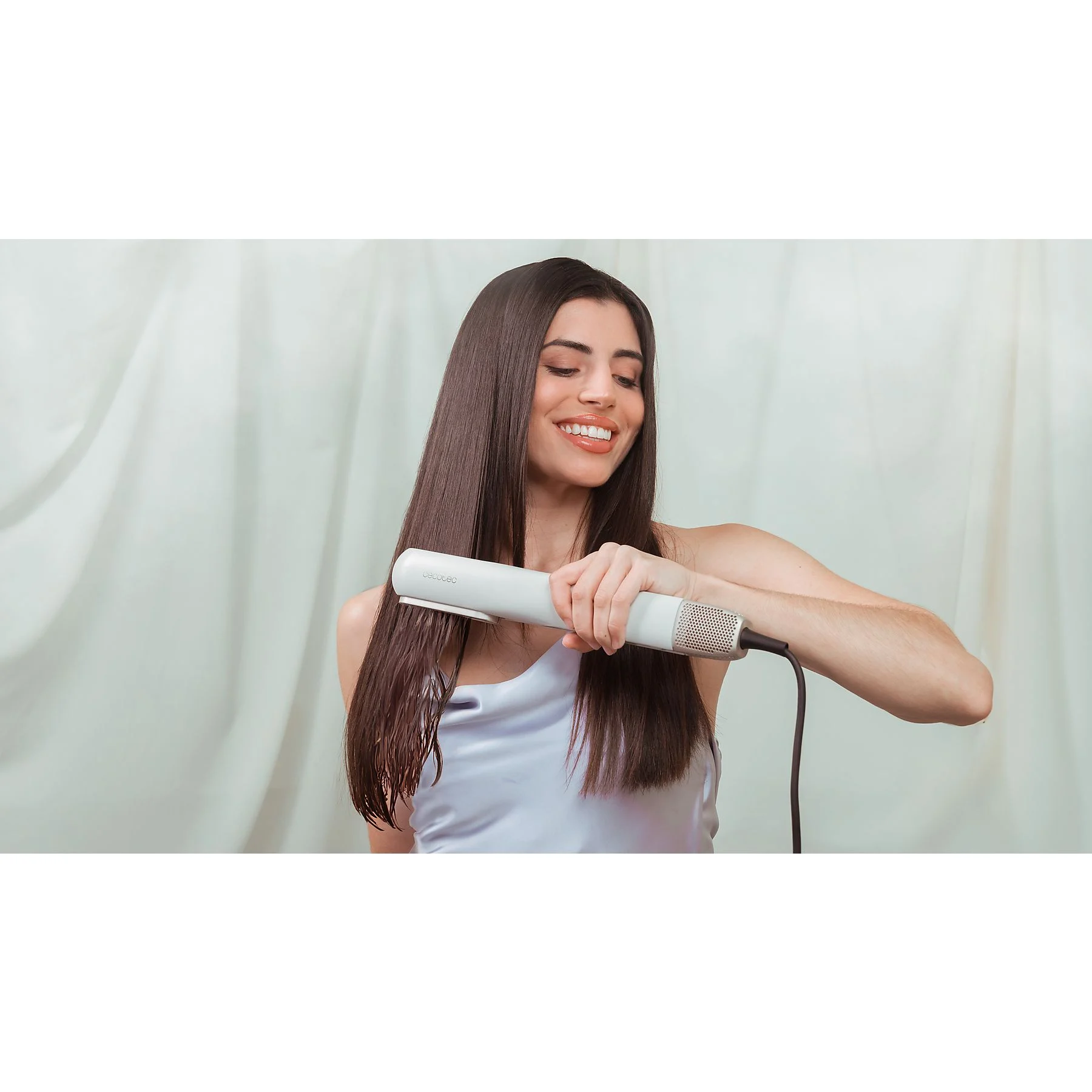 Kobieta używa prostownicy do swoich długich, ciemnych włosów. Uśmiecha się, trzymając białą prostownicę. Jasne tło z zasłonami.