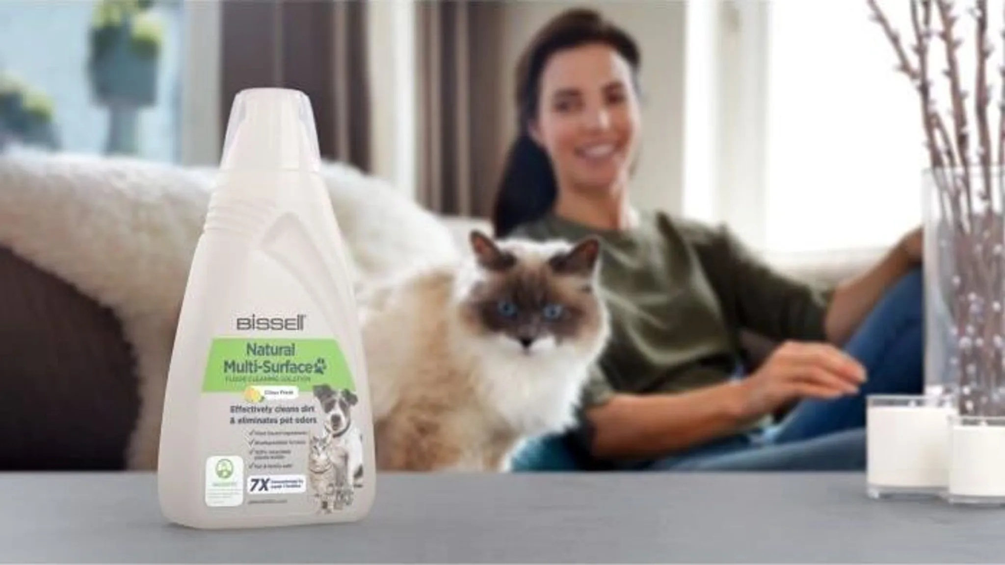 Kobieta siedzi z kotem. Z przodu butelka płynu czyszczącego. Świece i kwiaty w tle.