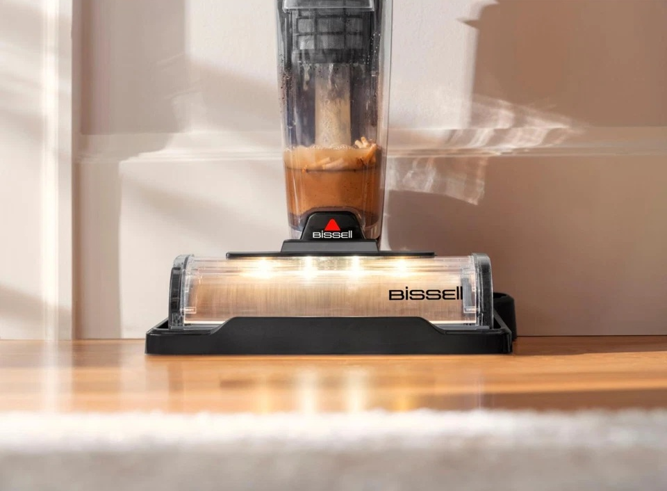 Na drewnianej podłodze pokazano środek do czyszczenia podłóg Bissell. Odkurzacz ma przezroczysty pojemnik z brązowym płynem. Maszyna jest czarna.