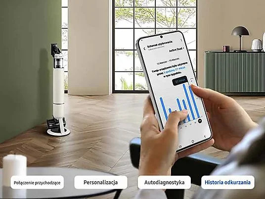 Osoba trzyma telefon z wyświetlonymi statystykami użytkowania. Odkurzacz stoi obok. W tle widać okno i meble.