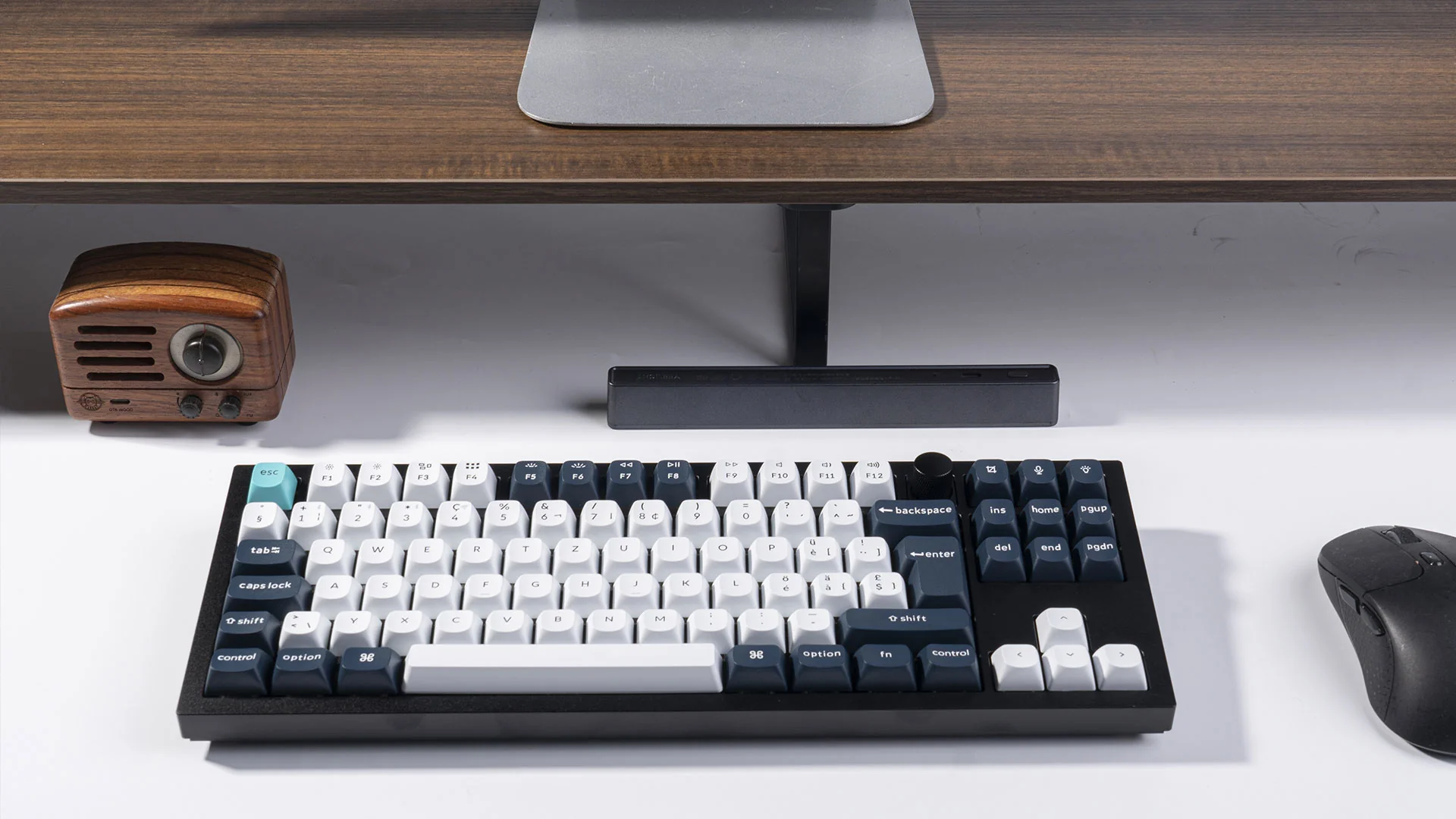 Eine mechanische Tastatur mit weißen und schwarzen Tasten liegt auf einem weißen Schreibtisch. Ein Holzradio und eine Maus sind ebenfalls vorhanden.