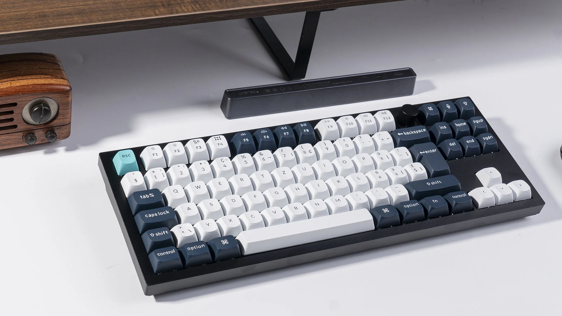 Eine schwarz-weiße Tastatur liegt auf einer weißen Oberfläche. Oben links befindet sich ein kleines Holzradio und darüber eine schwarze Leiste.