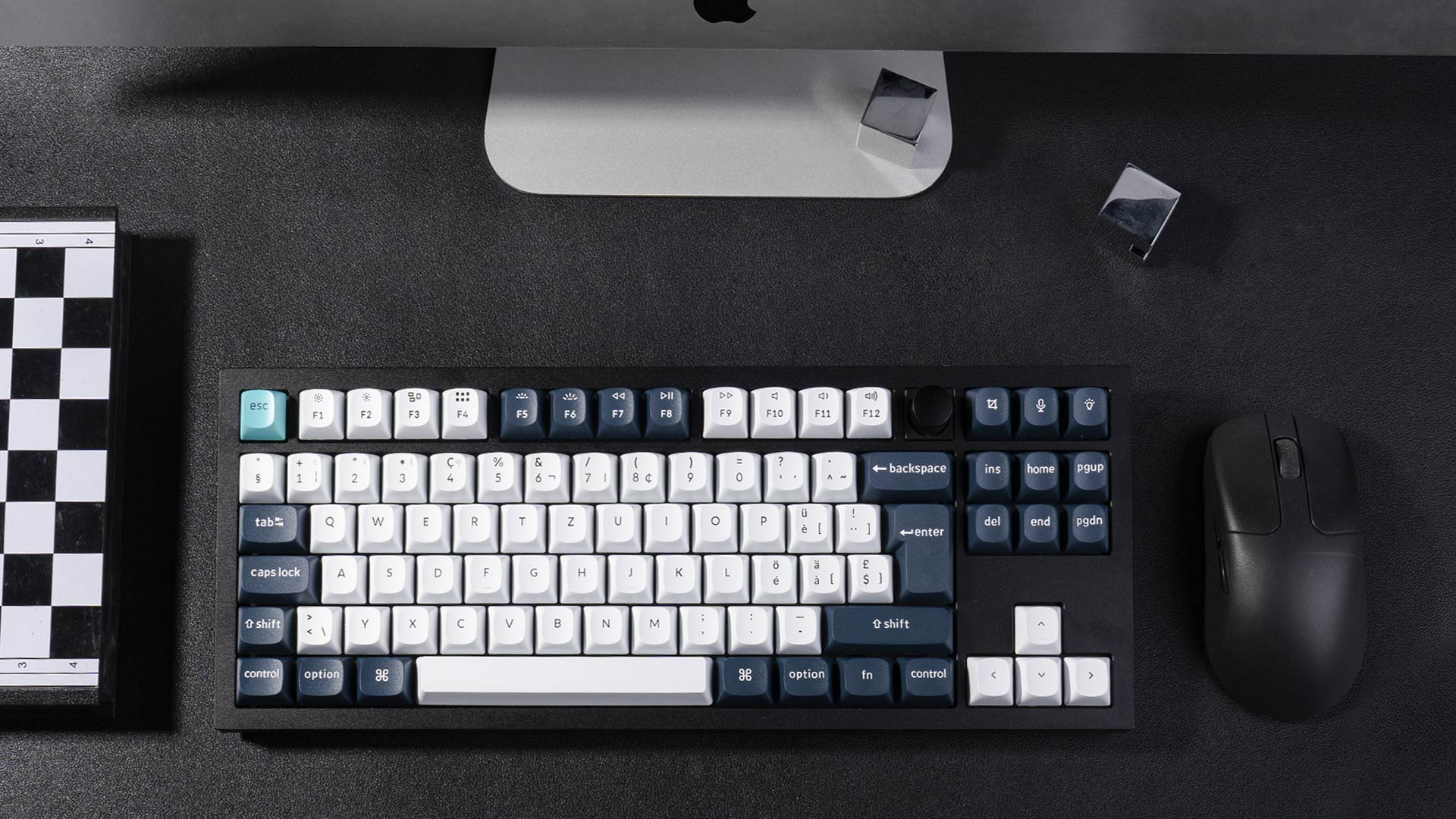Draufsicht auf Tastatur, Maus und Schachbrett auf schwarzer Oberfläche, mit Computermonitor im Hintergrund.
