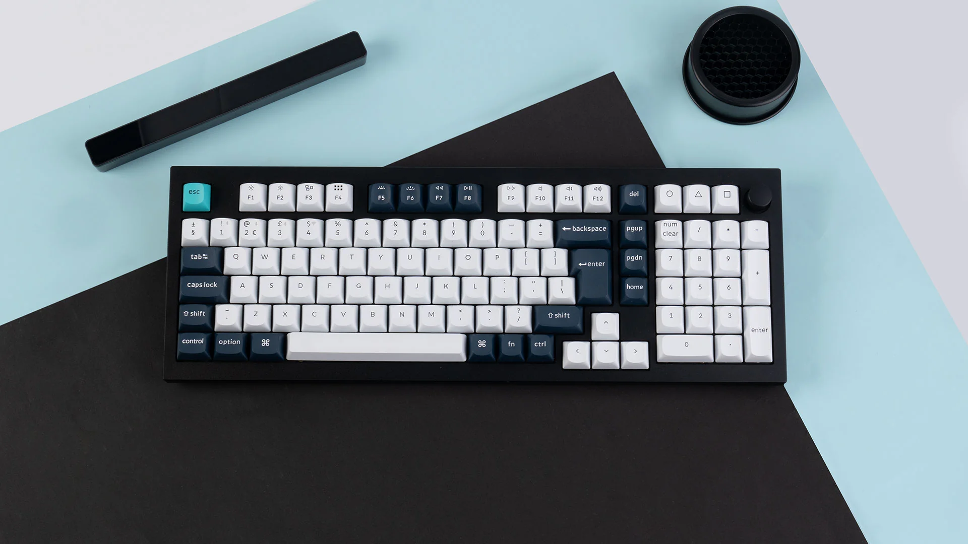 Eine schwarze Tastatur mit weißen und blauen Tasten liegt auf einer schwarz-hellblauen Oberfläche. Es gibt einen schwarzen Balken und ein rundes Objekt.