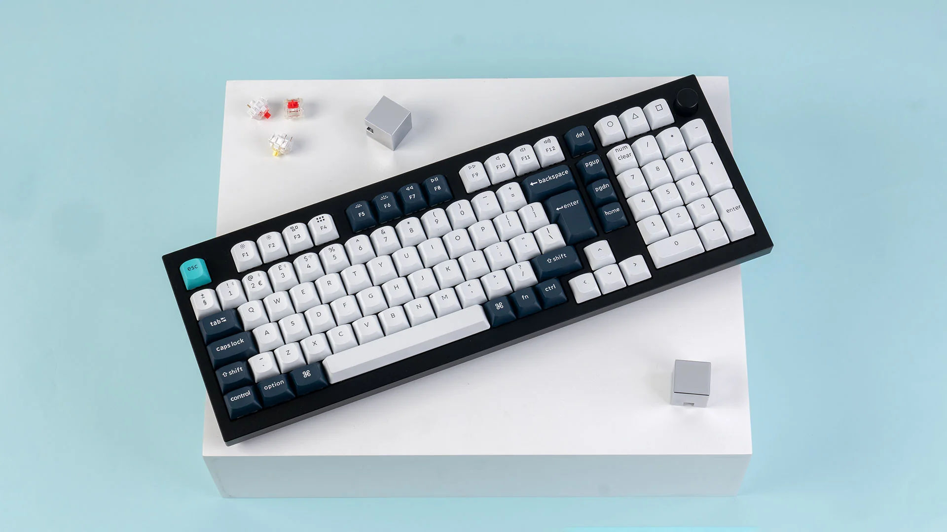 Eine schwarz-weiße mechanische Tastatur auf einem weißen Block, mit hellblauem Hintergrund. Drei Schalter befinden sich oben links.