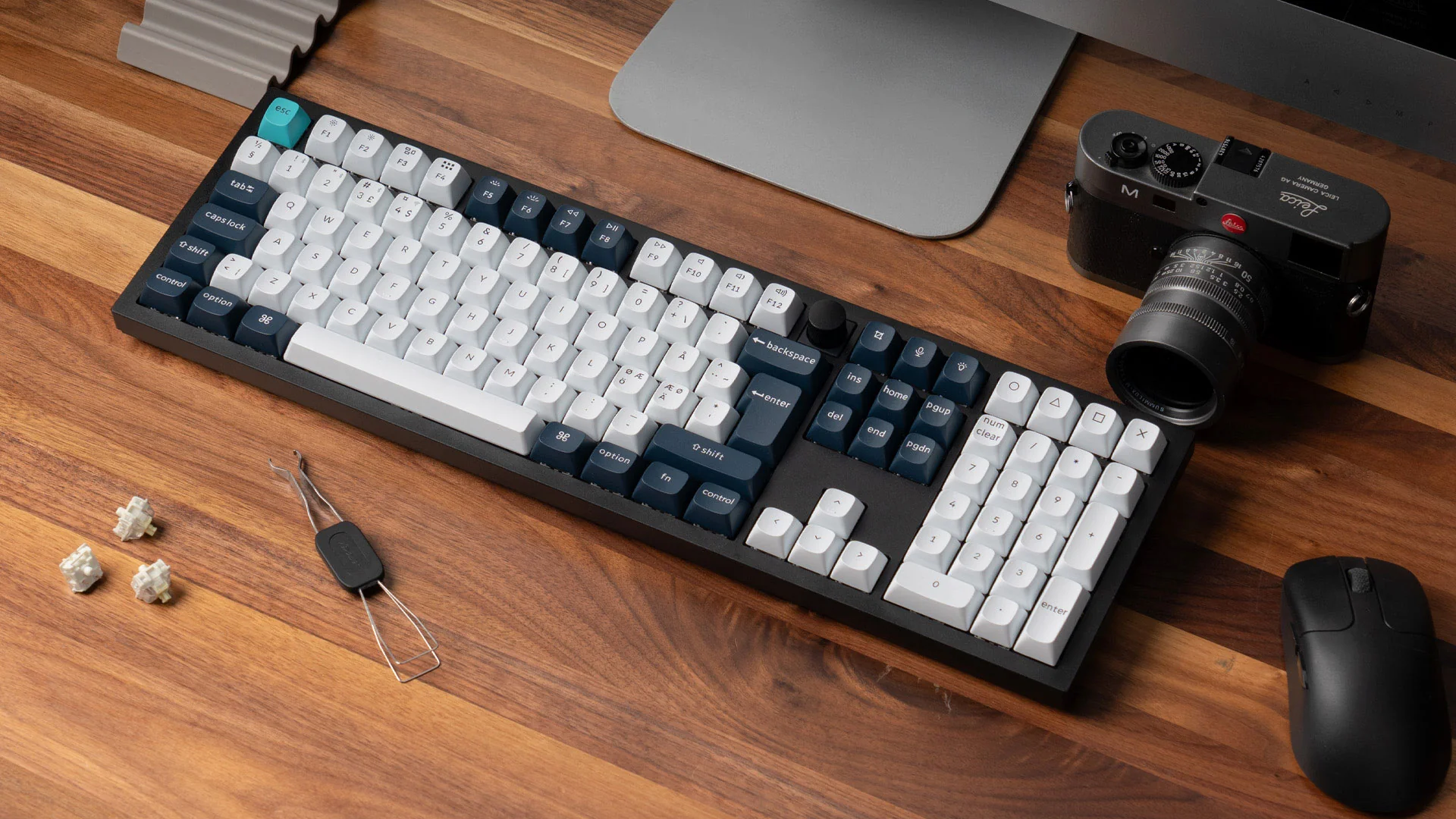 Eine Tastatur mit weißen und dunkelblauen Tasten steht auf einem Holztisch. Eine Kamera, eine Maus und eine Pinzette befinden sich in der Nähe.