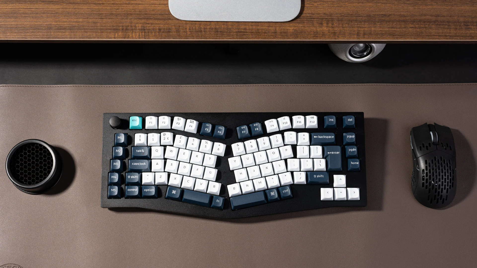 Eine ergonomische geteilte Tastatur, eine Maus und ein Lautsprecher befinden sich auf einer braunen Schreibtischunterlage.