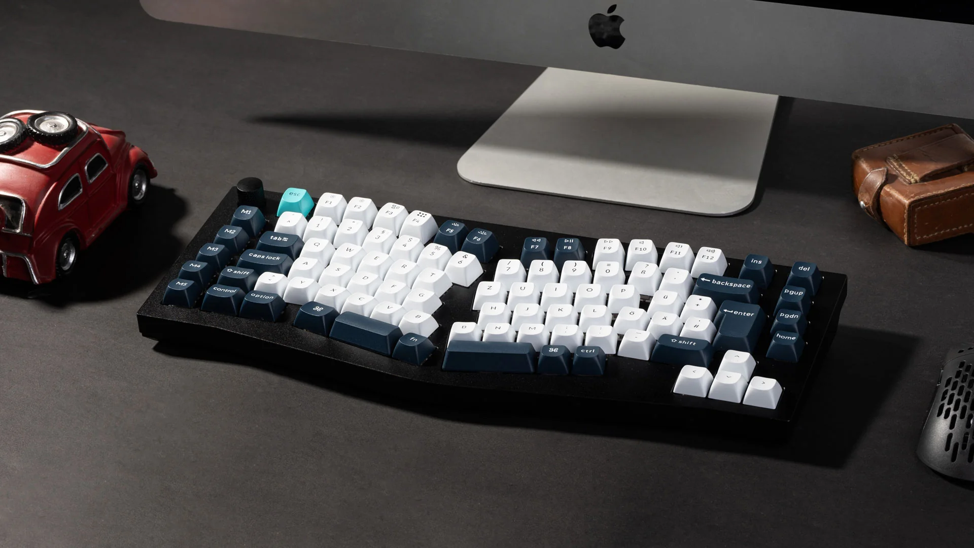 Eine schwarze mechanische Tastatur mit weißen und dunkelblauen Tasten, ein rotes Spielzeugauto und ein Apple-Monitor.