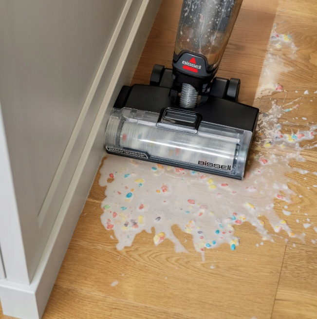 Odkurzacz Bissell sprząta rozlaną mieszankę kolorowych płatków śniadaniowych i mleka na drewnianej podłodze.