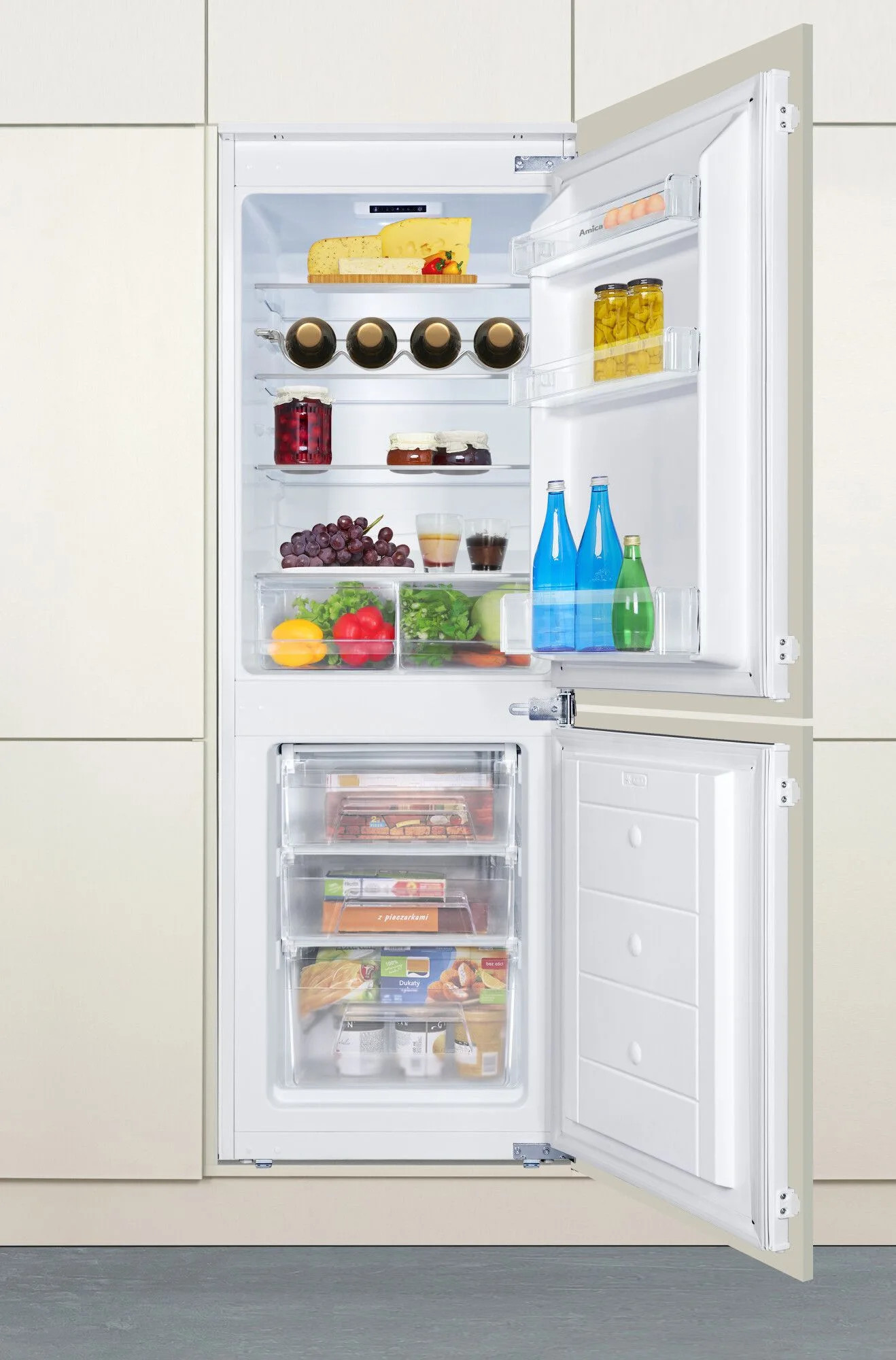 Otwarta lodówka z jedzeniem w środku. Widoczne są sery, słoiki, butelki, winogrona, warzywa i mrożonki. Lodówka jest biała.