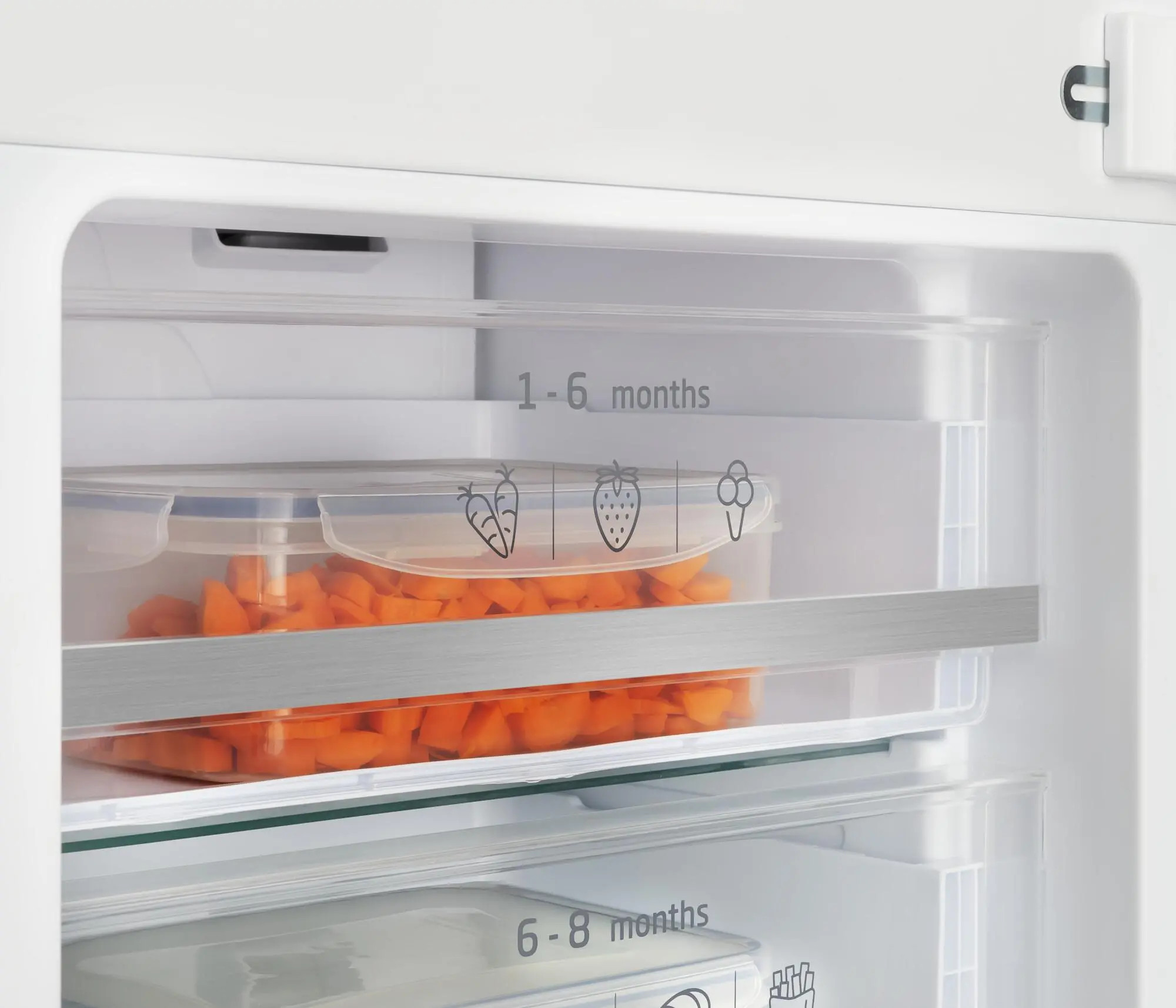 Wewnątrz lodówki widoczny pojemnik z pokrojoną marchewką. Etykiety wskazują czasy przechowywania. Lodówka jest biała.