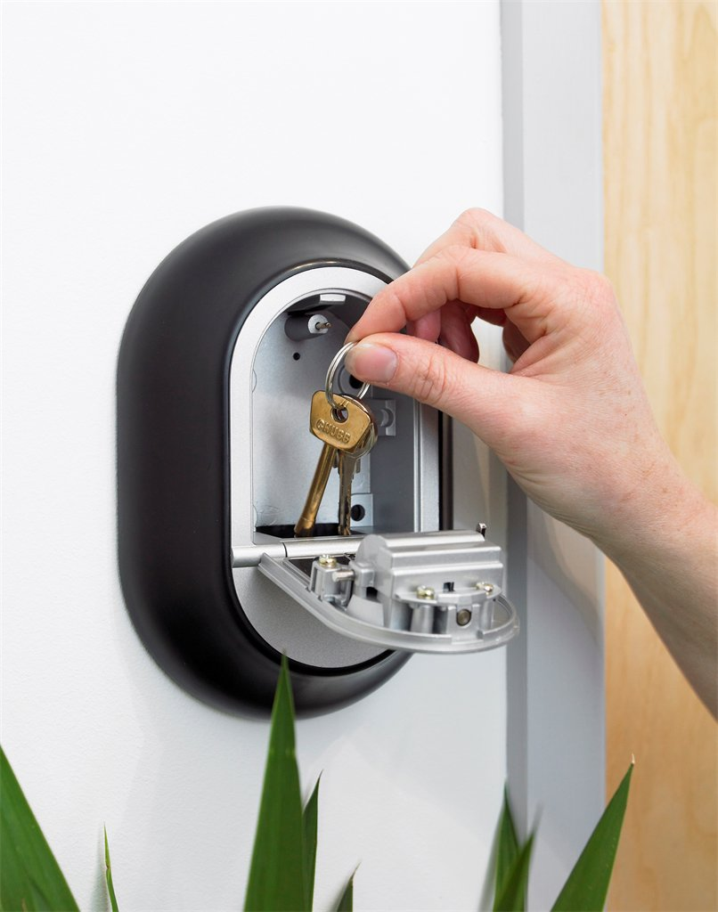 Eine Hand entnimmt Schlüssel aus einem an der Wand befestigten Schlüsselsafe. Der Safe ist schwarz, mit silbernem Innenraum und einer Flügeltür. Die Schlüssel sind goldfarben.