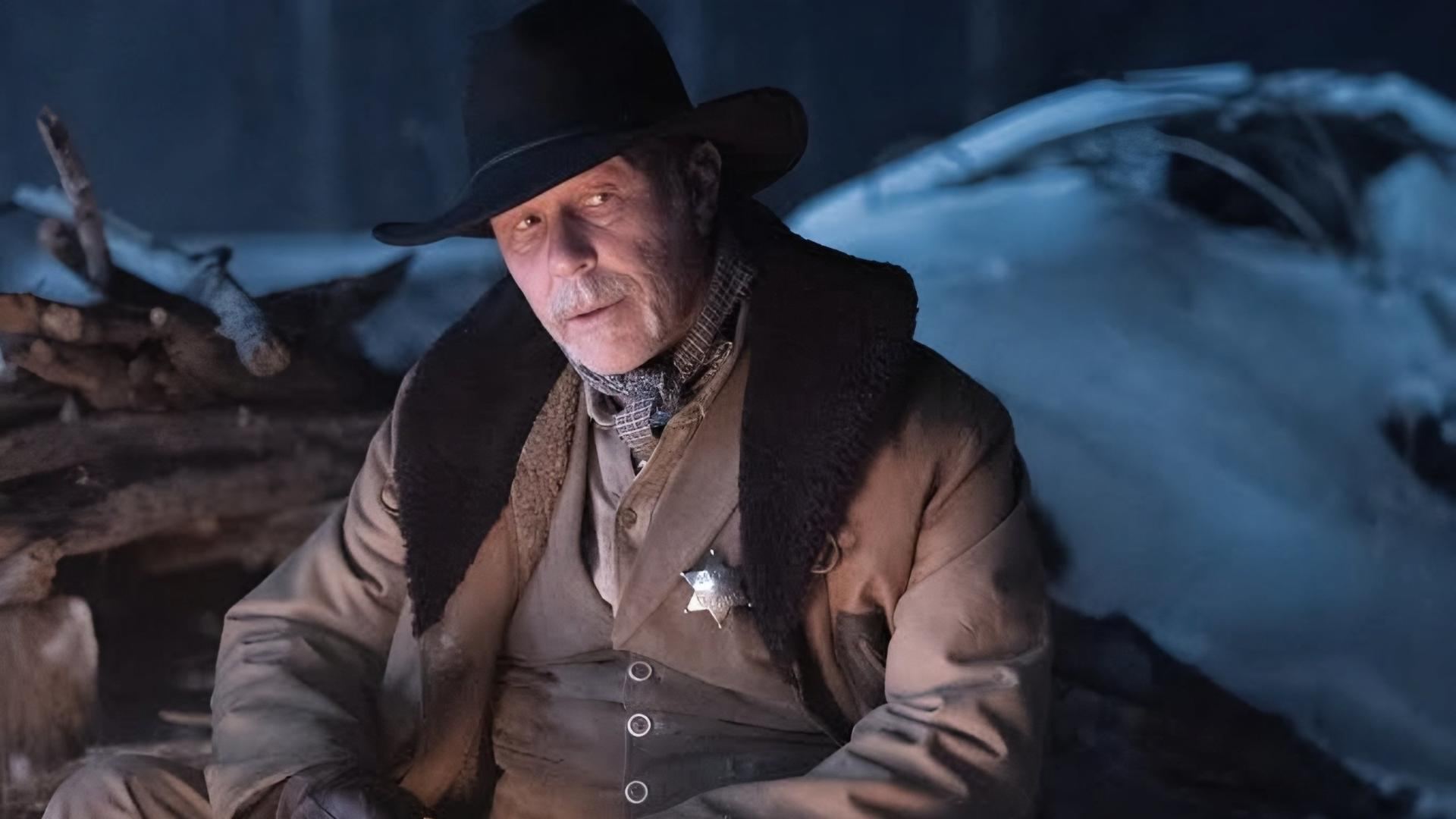 Ein älterer Mann mit Cowboyhut und Mantel, der einen Sheriffstern trägt. Verschneiter Hintergrund mit Baumstämmen.