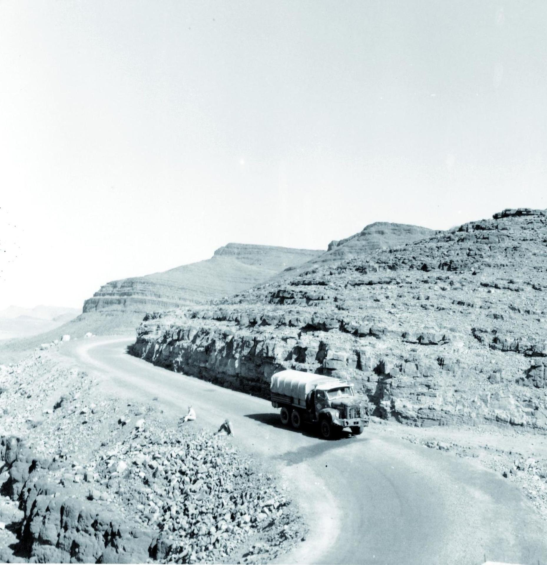Ein Lastwagen fährt auf einer kurvenreichen Straße in einer Wüstenlandschaft. Felsige Berge sind im Hintergrund. Eine Person sitzt am Straßenrand.