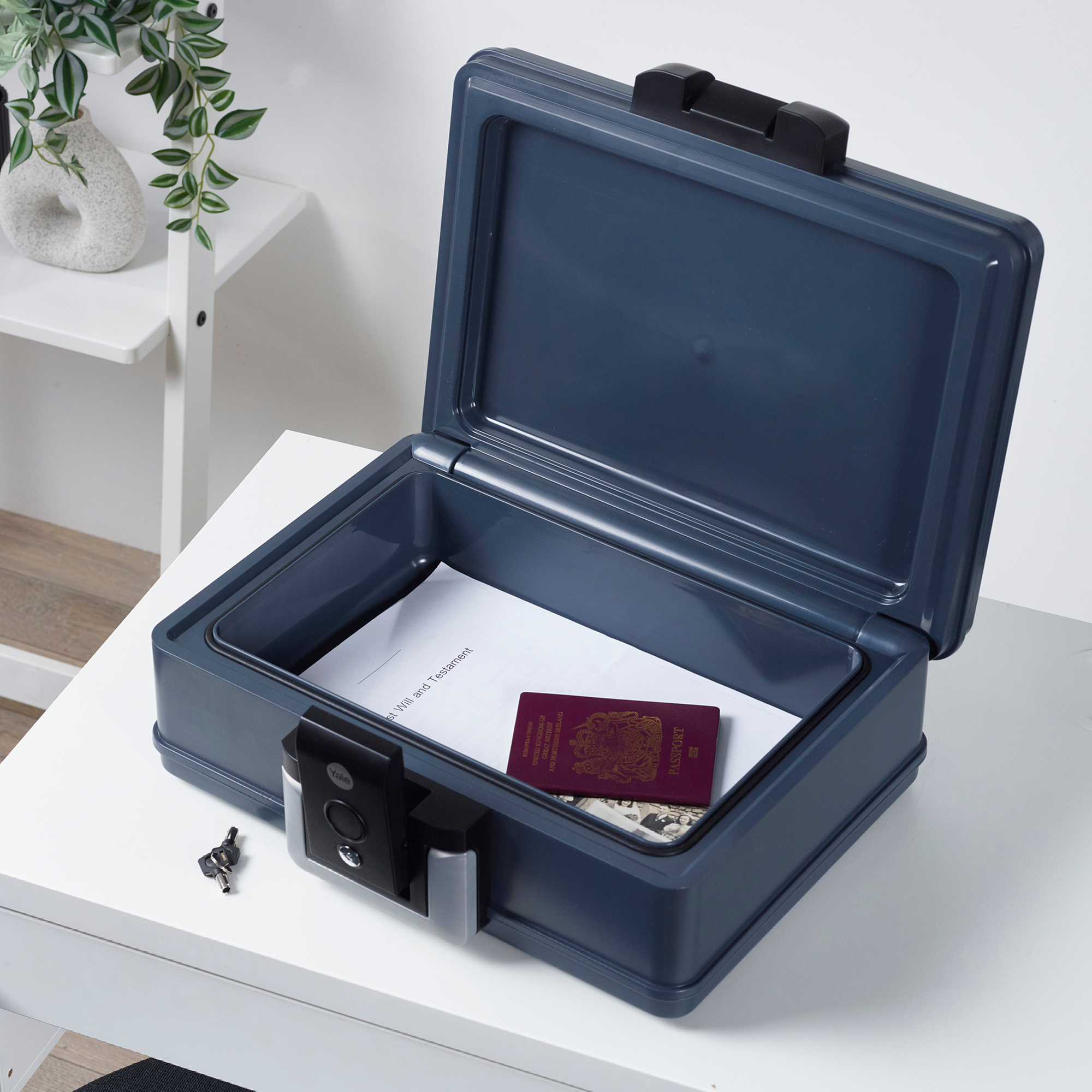Ein offener, dunkelblauer Safe enthält Dokumente und einen Reisepass. Schlüssel liegen auf dem Tisch. Im Hintergrund ein weißes Regal mit einer Pflanze.