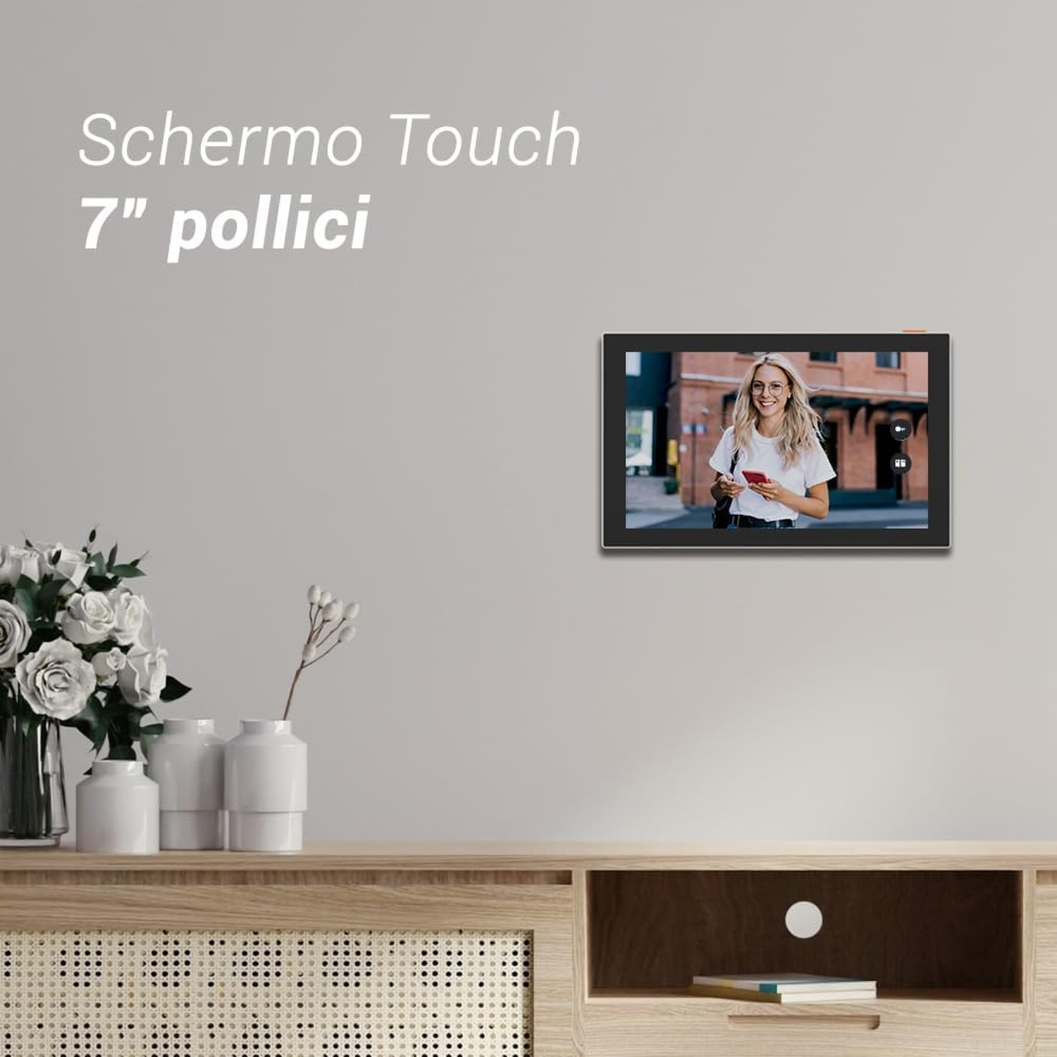 Un touchscreen montato a parete mostra una donna che tiene un telefono. Fiori bianchi su un mobile di legno.