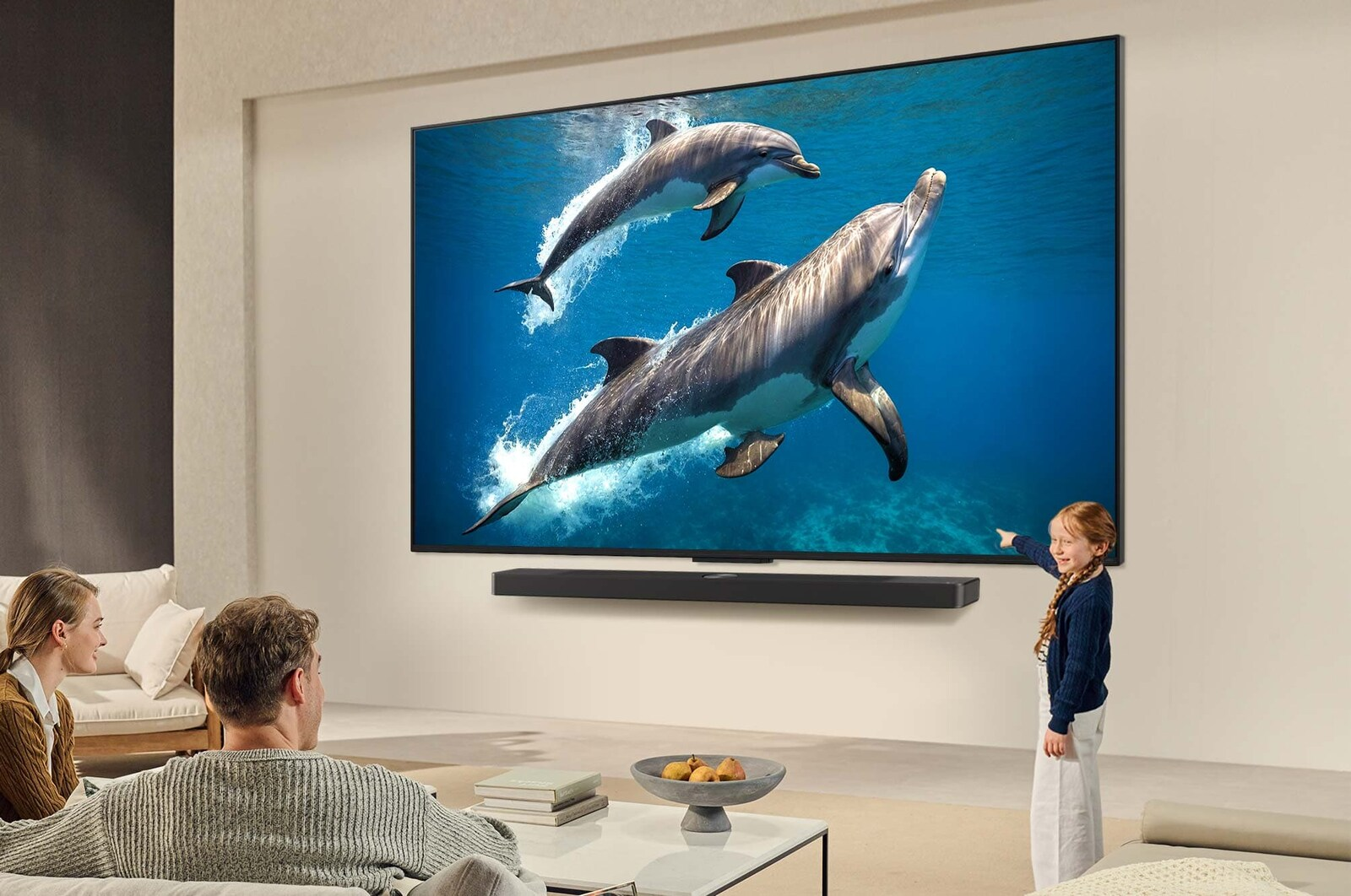 Familie schaut fern. Delphine schwimmen auf dem Bildschirm. Ein Mädchen zeigt auf den Fernseher. Eine Soundbar darunter.