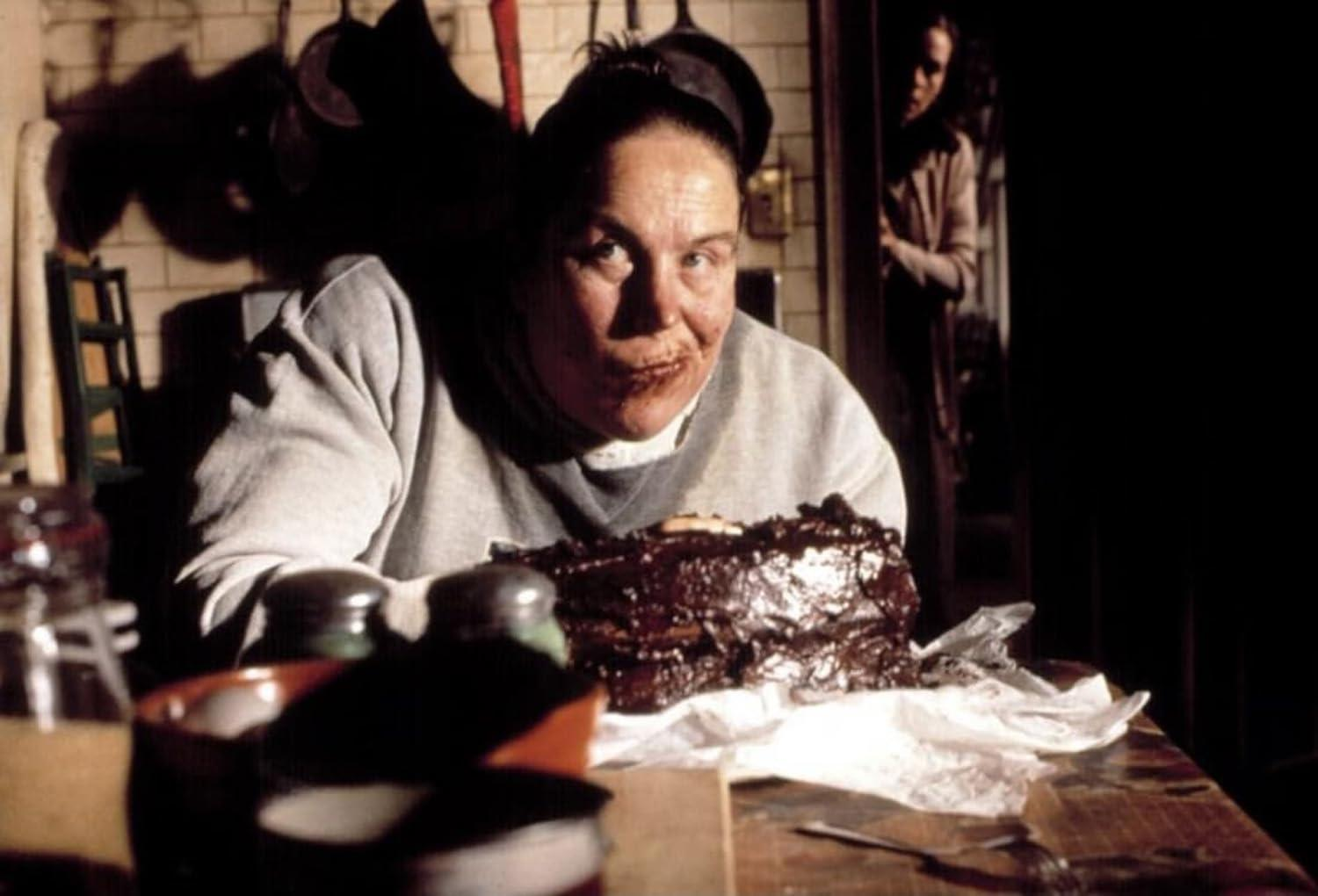 Eine Frau mit Schokolade im Mund sitzt hinter einem Schokoladenkuchen. Eine andere Frau steht in der Tür. Küchenutensilien hängen im Hintergrund.