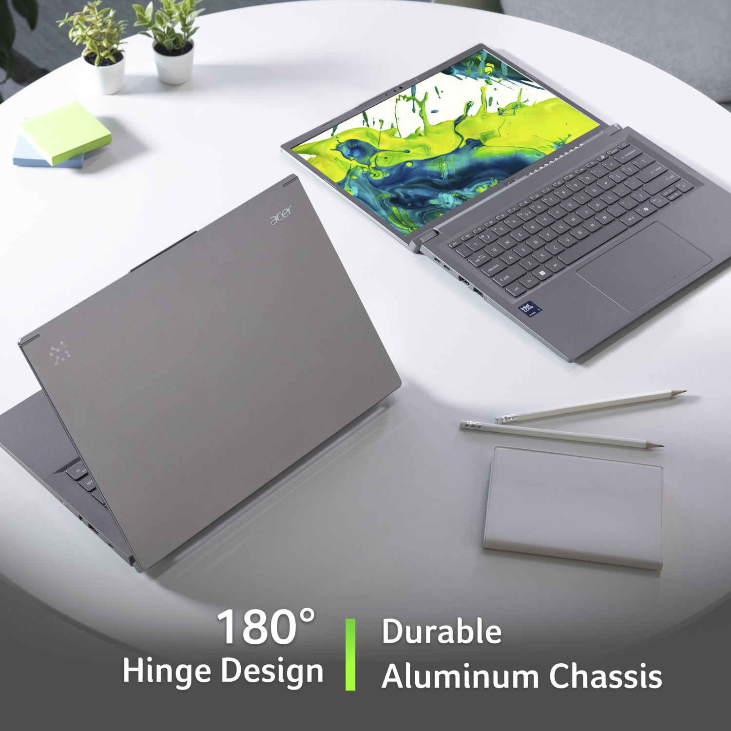 Two gray Acer laptops on a white table, one open displaying a colorful screen. Pencils, a pad, and plants are also present.