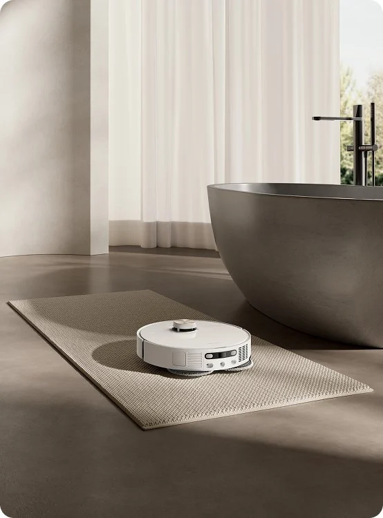 Egy fehér robotporszívó egy bézs szőnyegen. Mögötte egy szürke fürdőkád található. Fehér függönyök és ablak a háttérben.