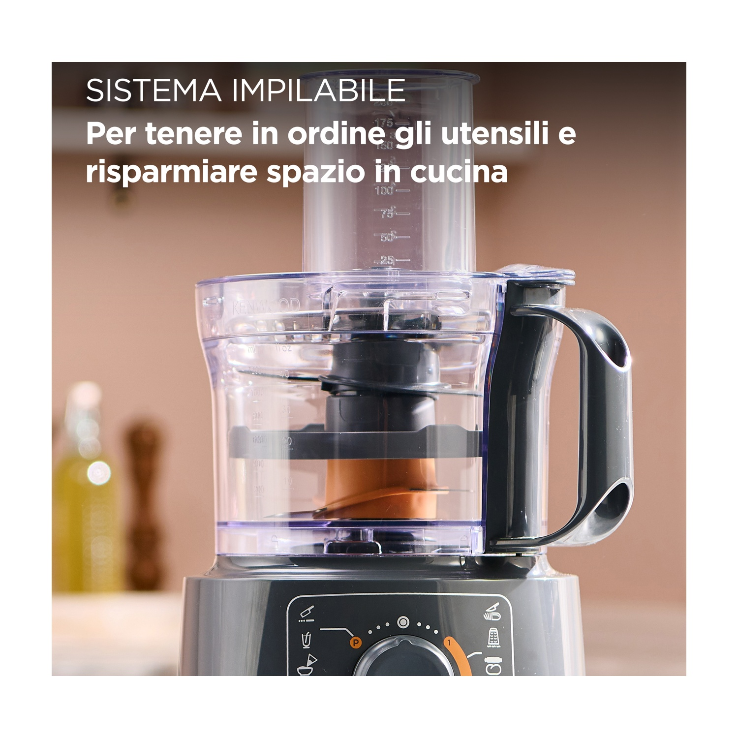 Un robot da cucina, ciotola e coperchio in plastica trasparente, manico nero. Lo sfondo è una scena di cucina sfocata con una bottiglia.