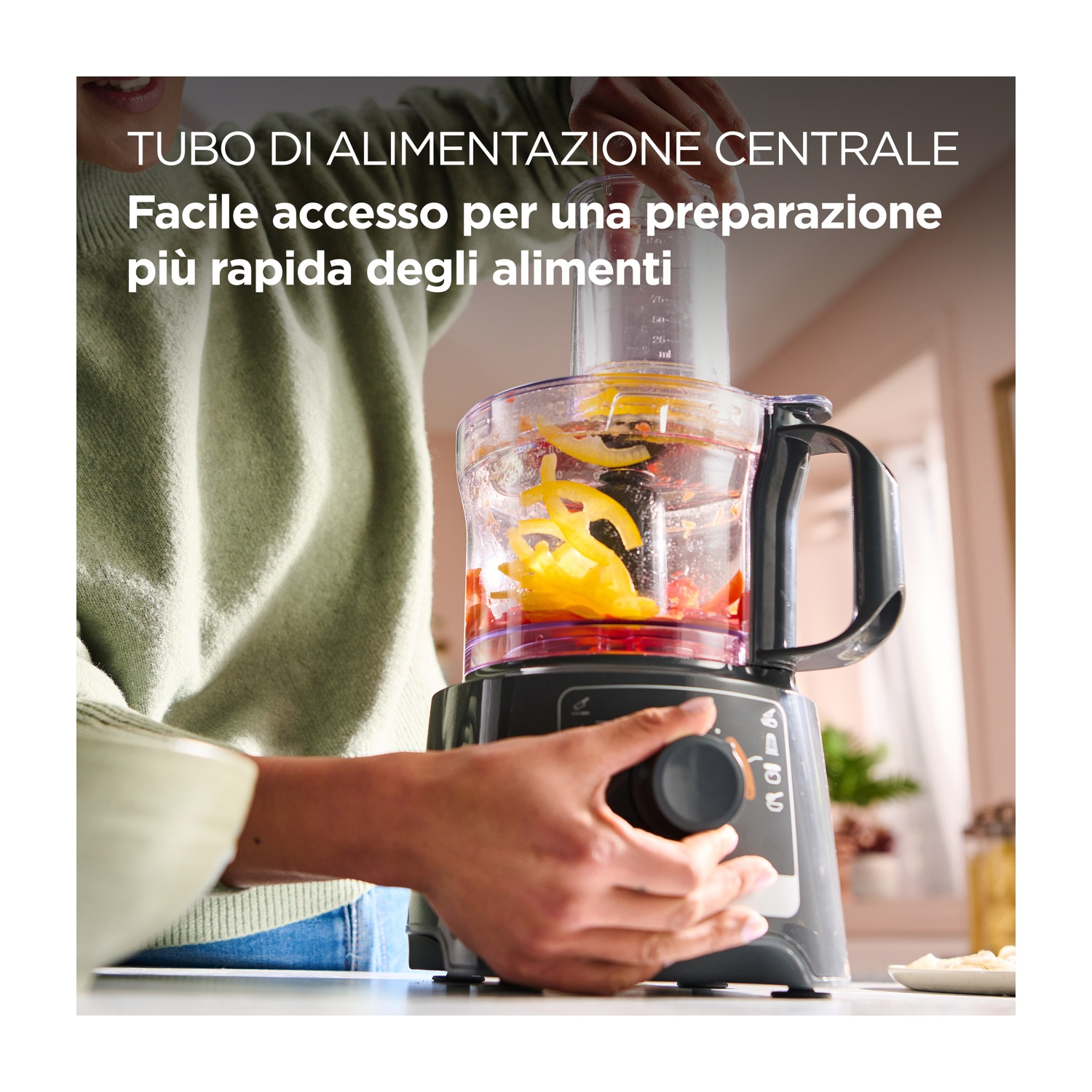 Una persona aziona un robot da cucina con verdure all'interno. La macchina è nera e grigia. La persona indossa un maglione verde.