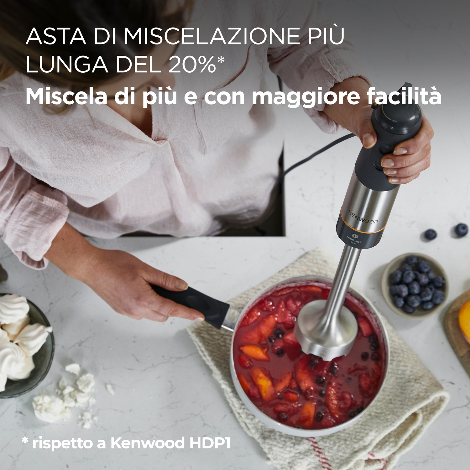 Una persona che mescola una miscela di frutta in una pentola con un Kenwood Triblade. Gli ingredienti includono pesche e mirtilli. Altri ingredienti sono sul lato.