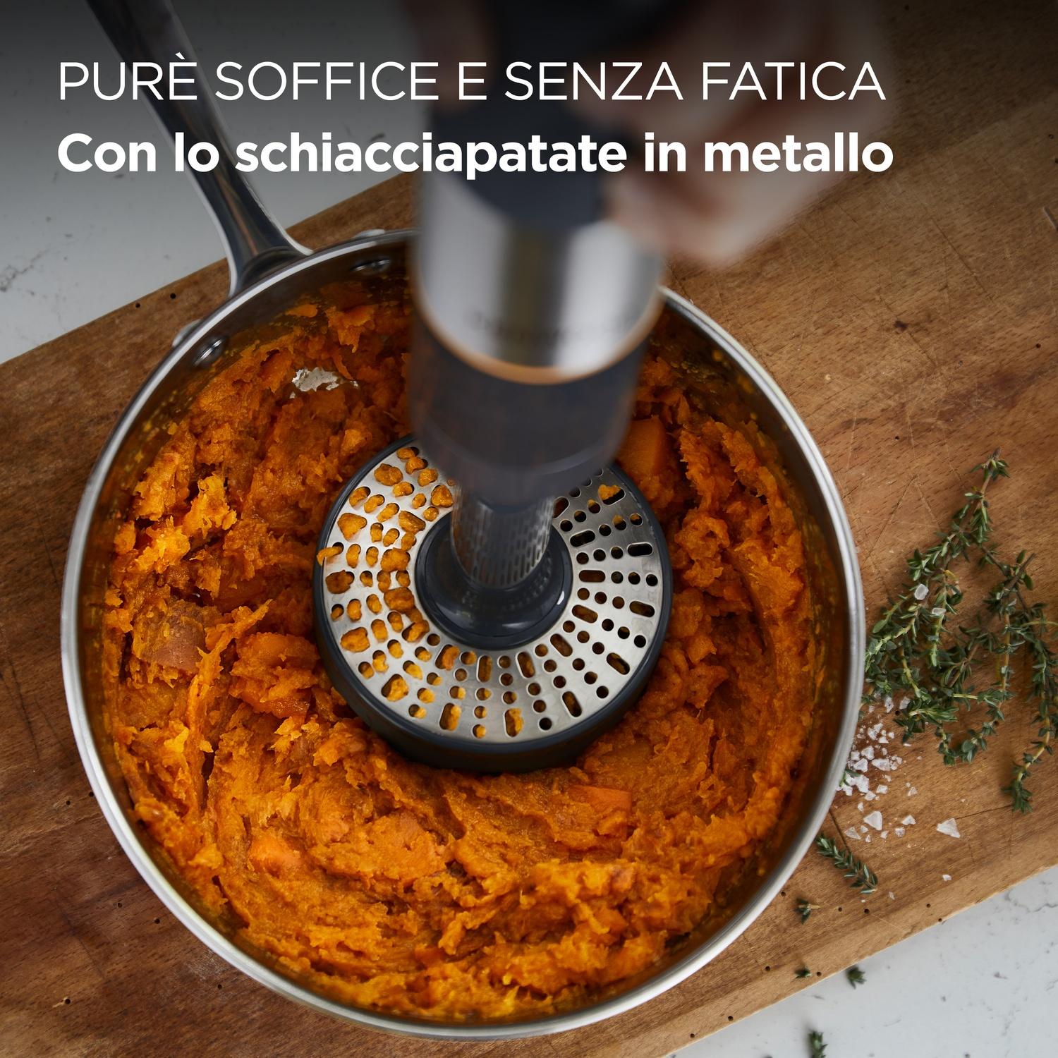 Un frullatore a immersione schiaccia cibo arancione in una pentola. La pentola è su un tagliere con erbe e sale.