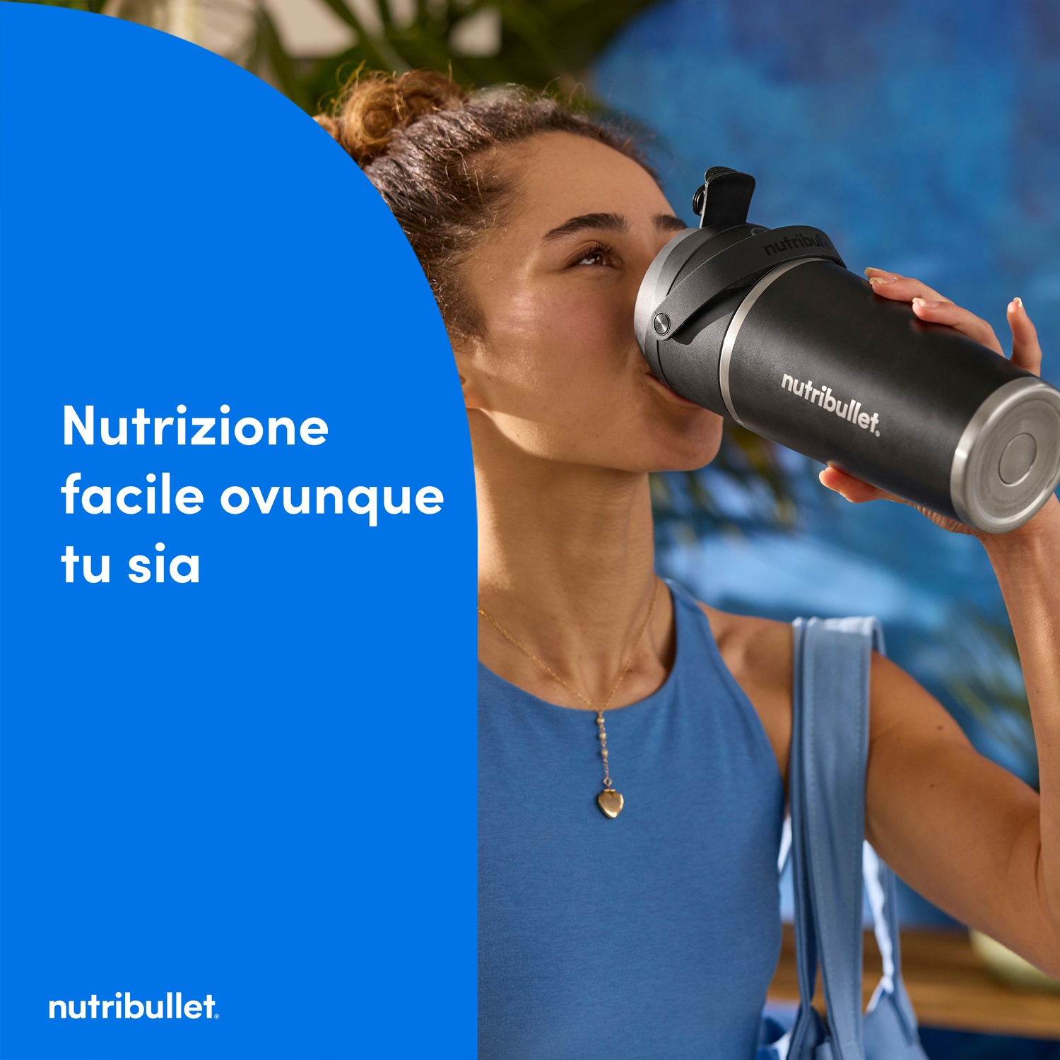 Una donna beve da uno shaker Nutribullet nero. Indossa abiti blu, una collana d'oro e una borsa azzurra sulla spalla.