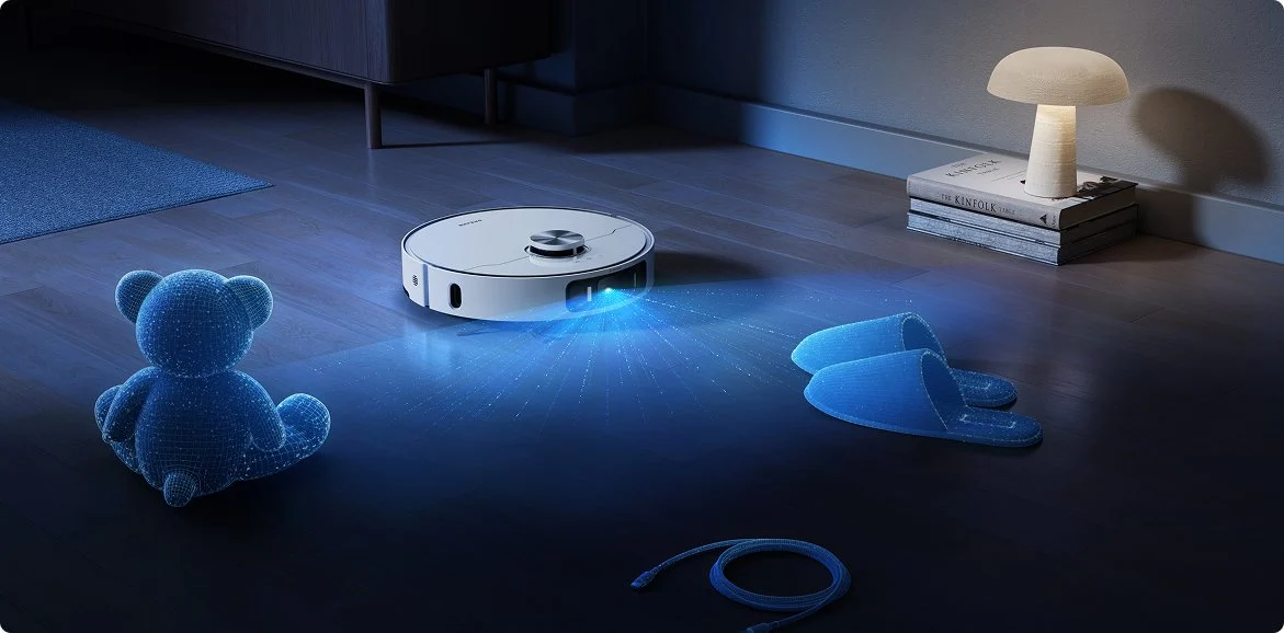 Egy fehér robotporszívó takarít egy szobát. Kék fénysugarak jönnek a porszívóból. Plüss maci és papucs látható.