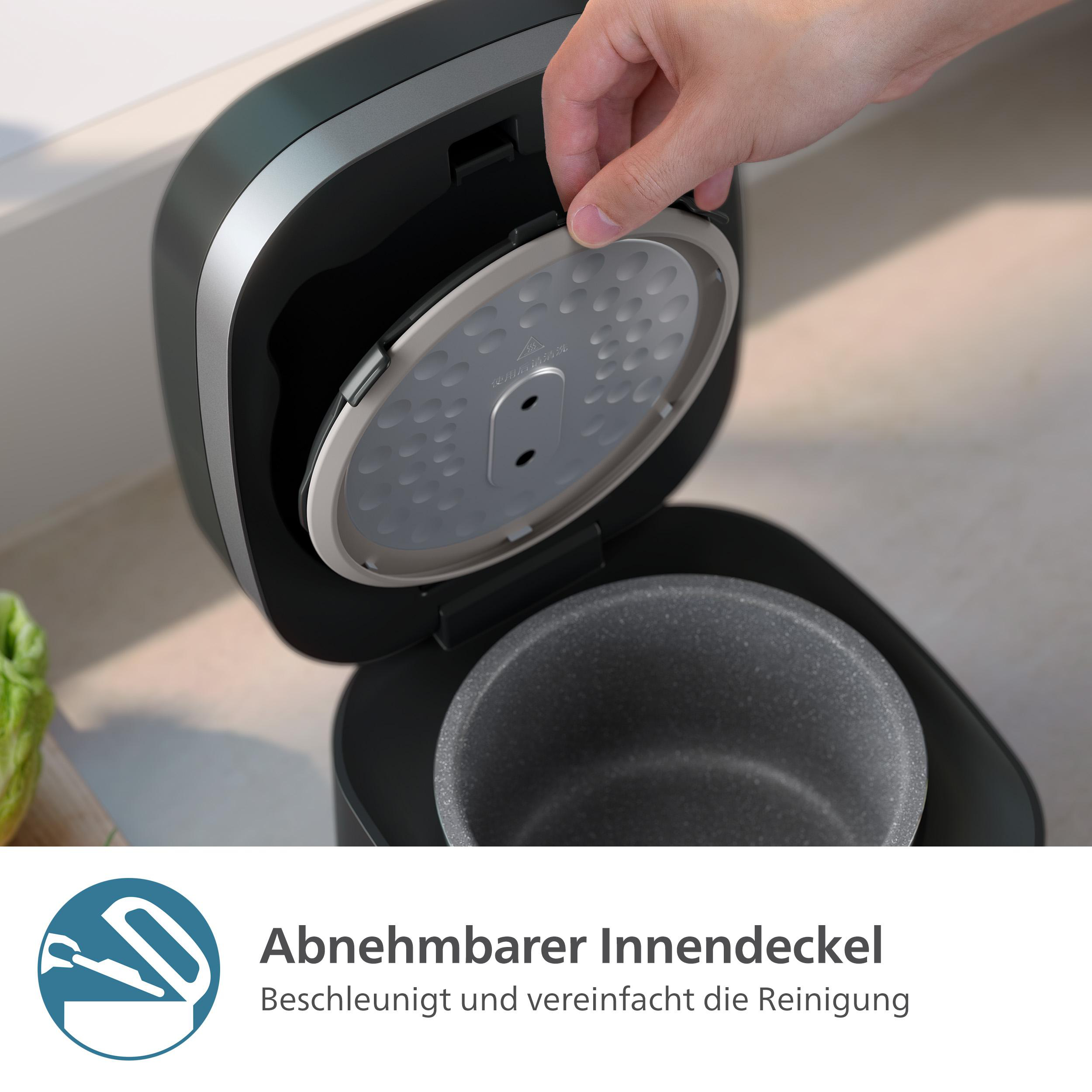 Eine Hand hebt den inneren Deckel eines schwarzen Reiskochers an. Der Deckel ist silberfarben und hat Löcher. Im Inneren befindet sich eine Schüssel.