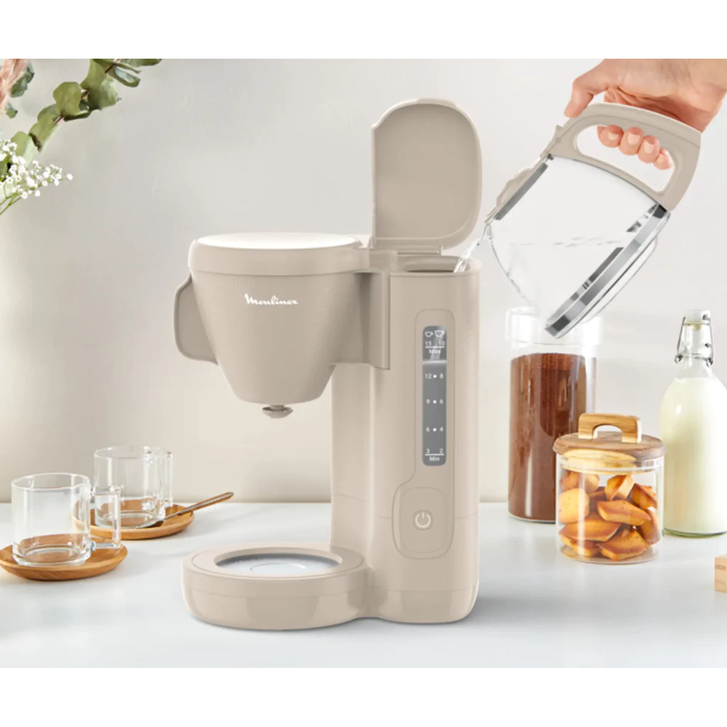 A person pouring water into a beige coffee maker. Transparent jars with coffee beans and cookies are visible. Two glasses sit on wooden saucers.