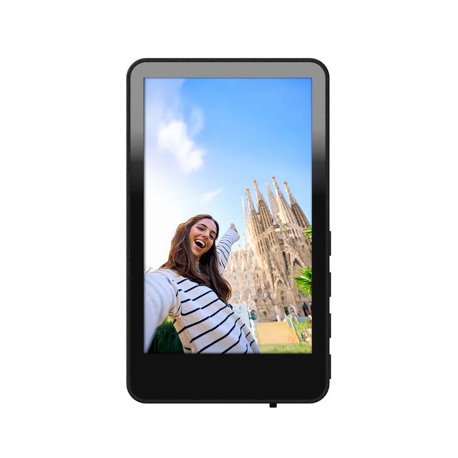 Eine Frau macht ein Selfie mit der Sagrada Familia im Hintergrund. Sie trägt ein gestreiftes Hemd, zeigt und lächelt. Klarer blauer Himmel.