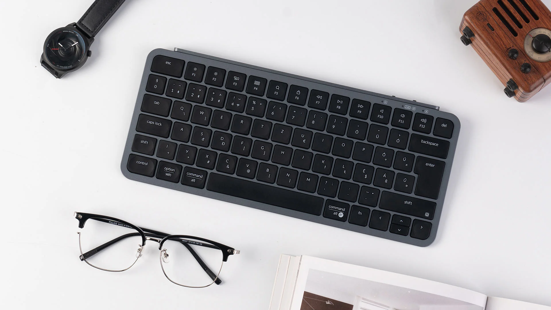 Eine graue Tastatur, eine Uhr, eine Brille und ein Radio liegen auf einer weißen Oberfläche. Ein Buch liegt in der Nähe der Brille.