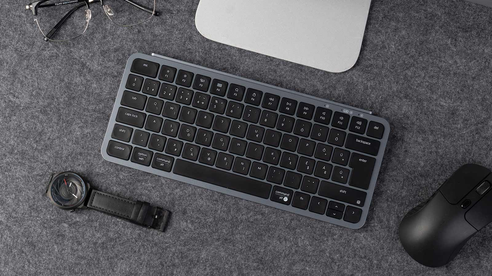 Draufsicht zeigt Tastatur, Maus und Uhr auf grauer Oberfläche. Brille links oben. Die Szene ist gut beleuchtet.