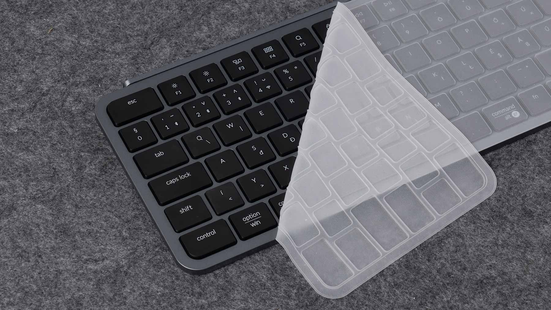 Eine dunkelgraue Tastatur mit schwarzen Tasten. Eine transparente Abdeckung bedeckt teilweise die Tastatur. Sie liegt auf einer grauen Oberfläche.